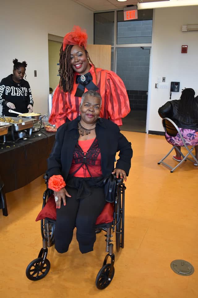 Two women in a room, one standing with colorful hair and the other sitting in a wheelchair with a red and black outfit.