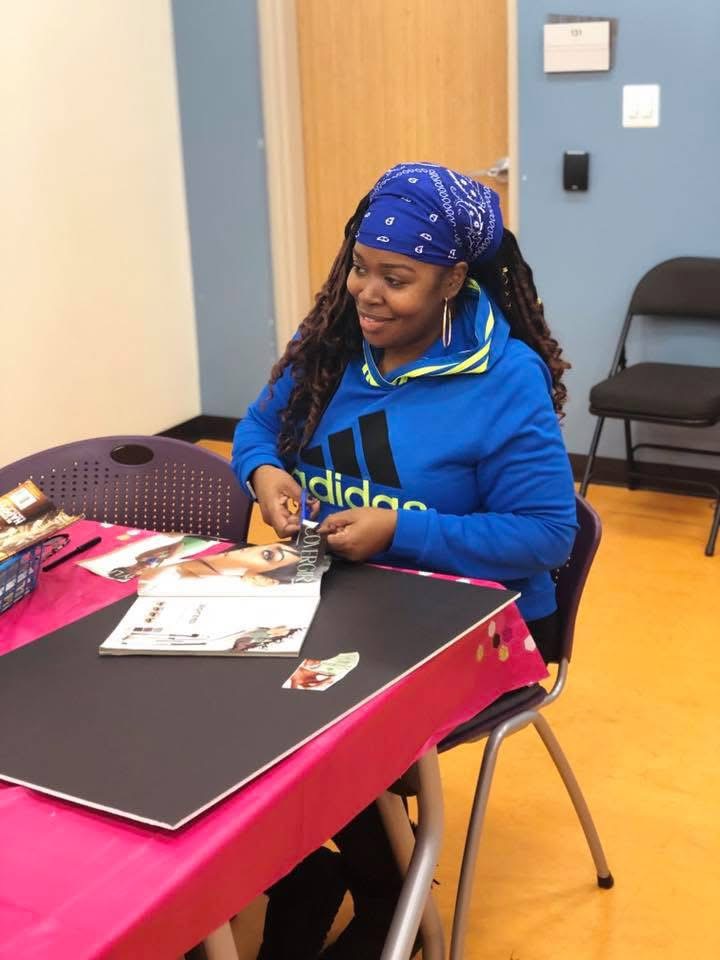 Girl sitting at table, wearing blue hoodie and bandana, smiling, with magazines and papers on table.