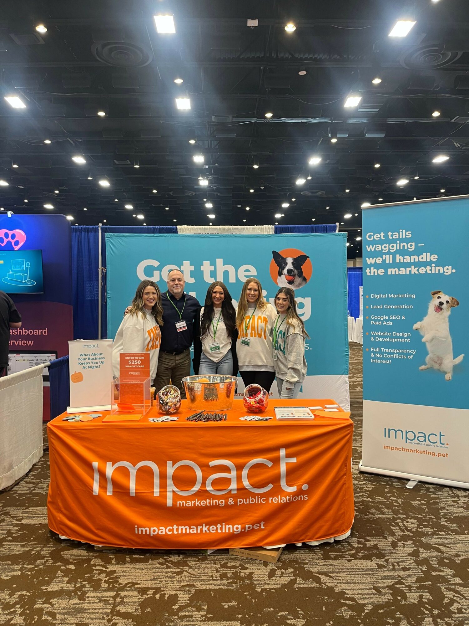 Four women standing behind an orange table with Impact branding at a conference booth, with banners in the background.