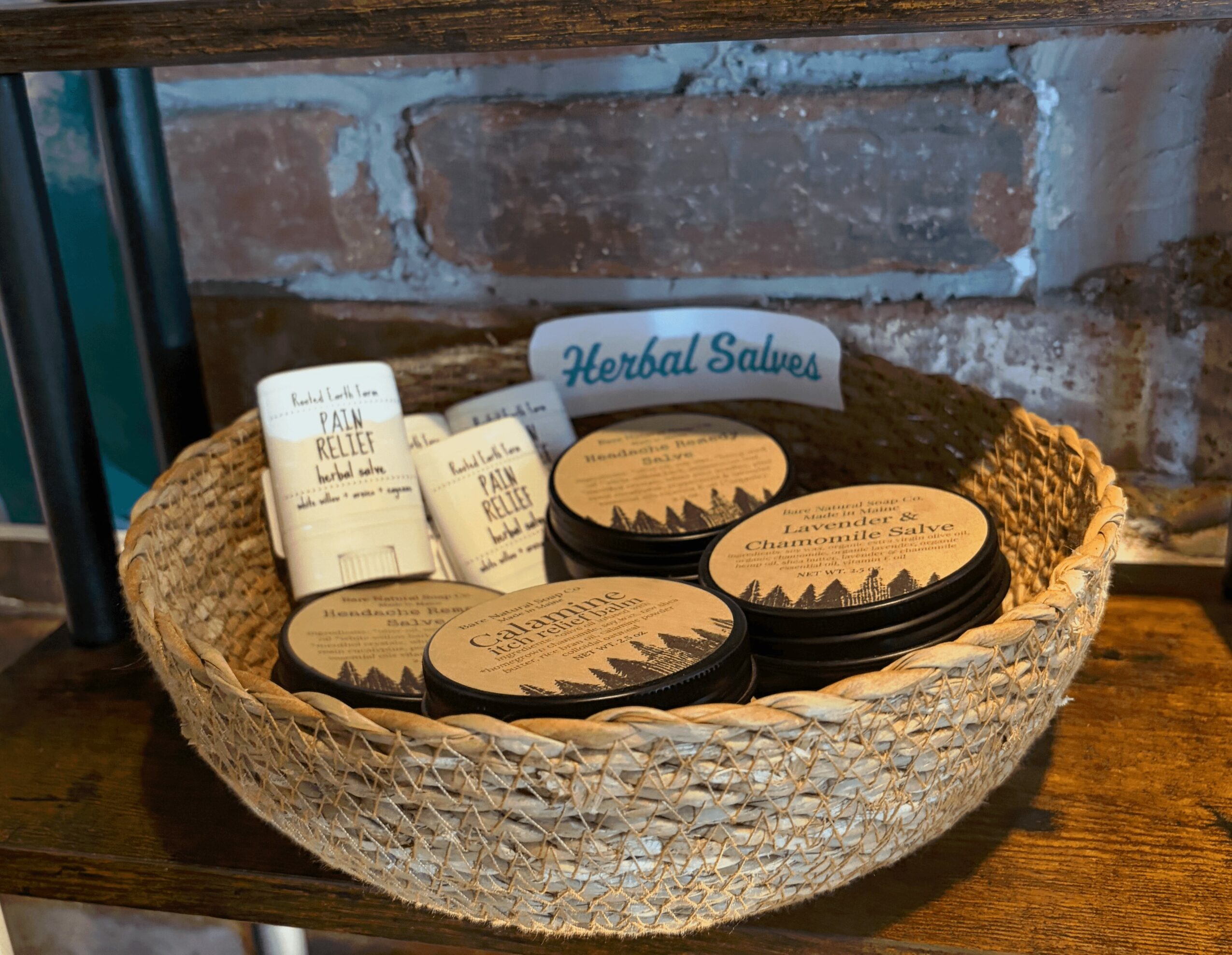 Basket with herbal salves and small containers on wooden surface against brick wall.