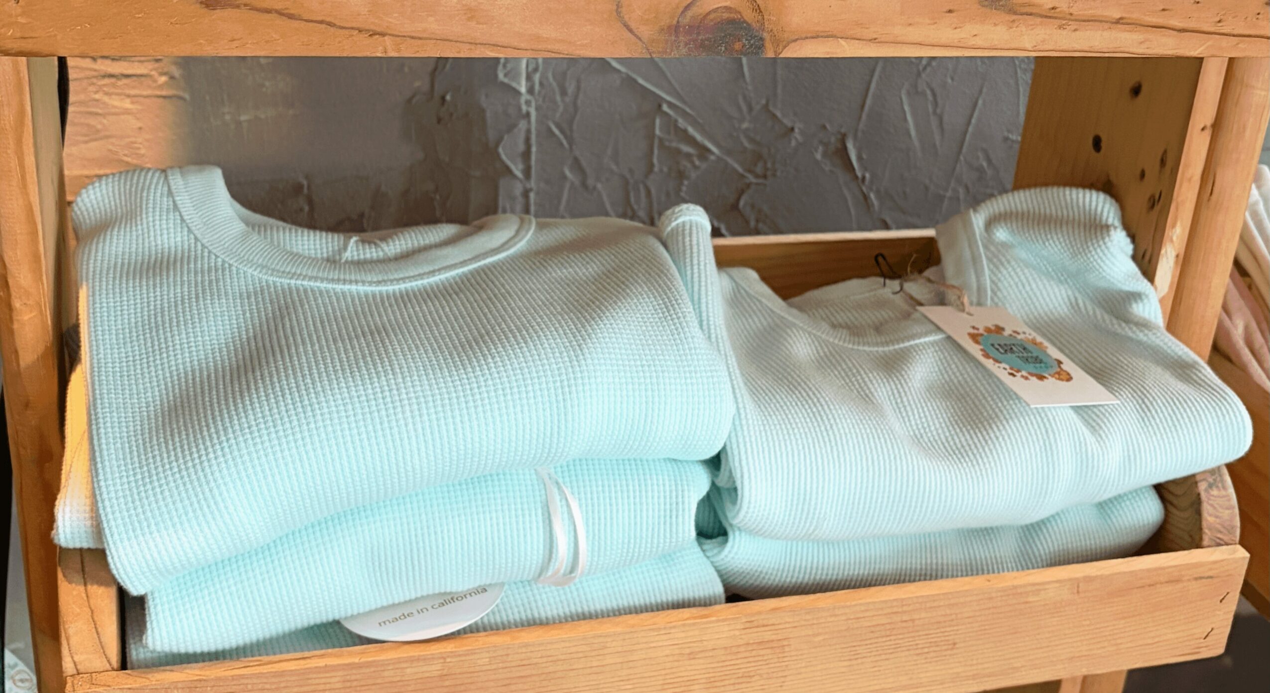Two folded light blue sweatshirts on a wooden shelf, one with a tag attached.