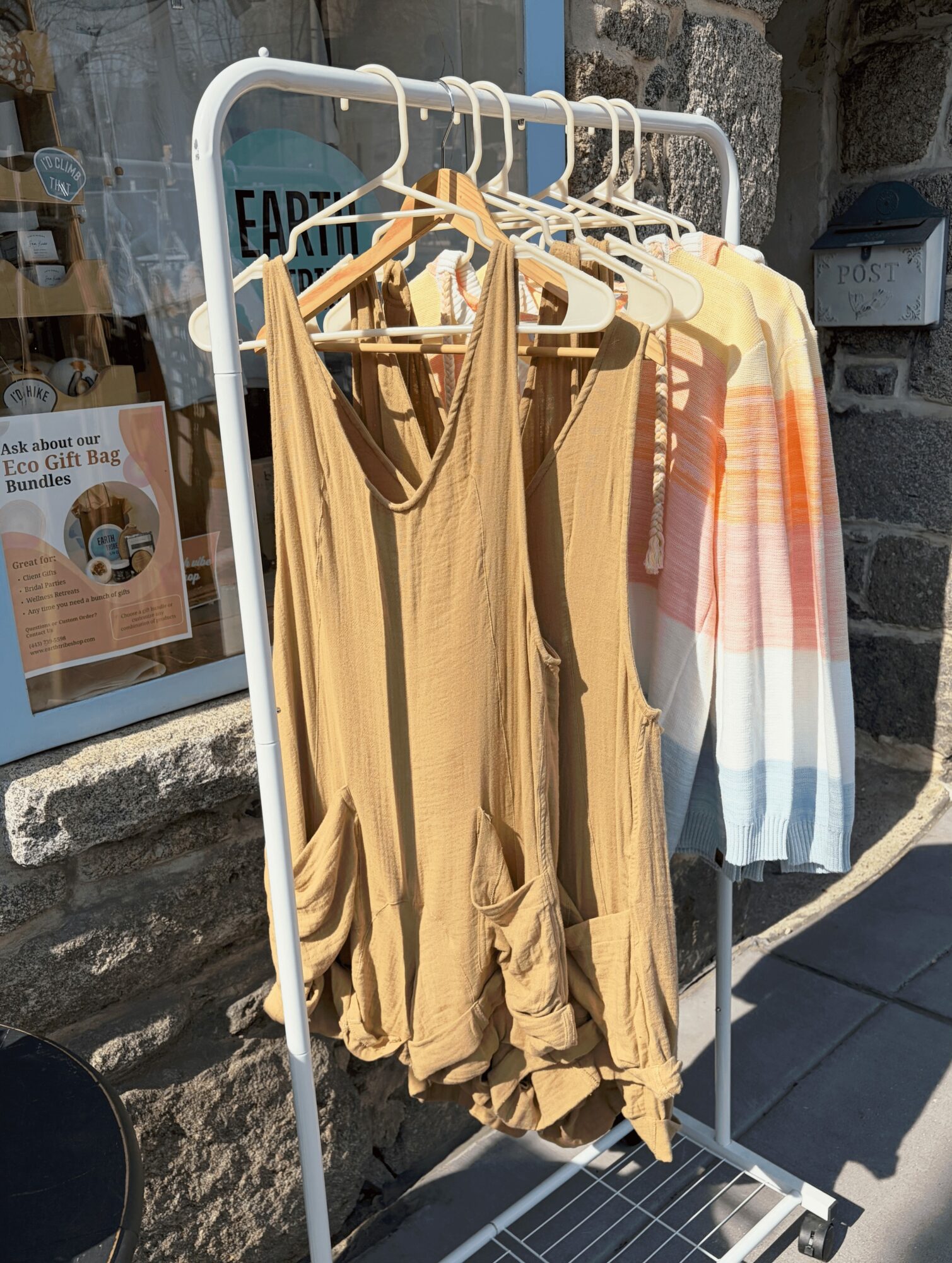 Clothing rack with beige and multicolored garments hanging outside a building with stone wall.