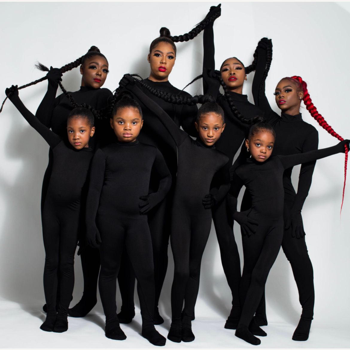 Eight women and girls in black outfits with braided hair, posing against a white background.