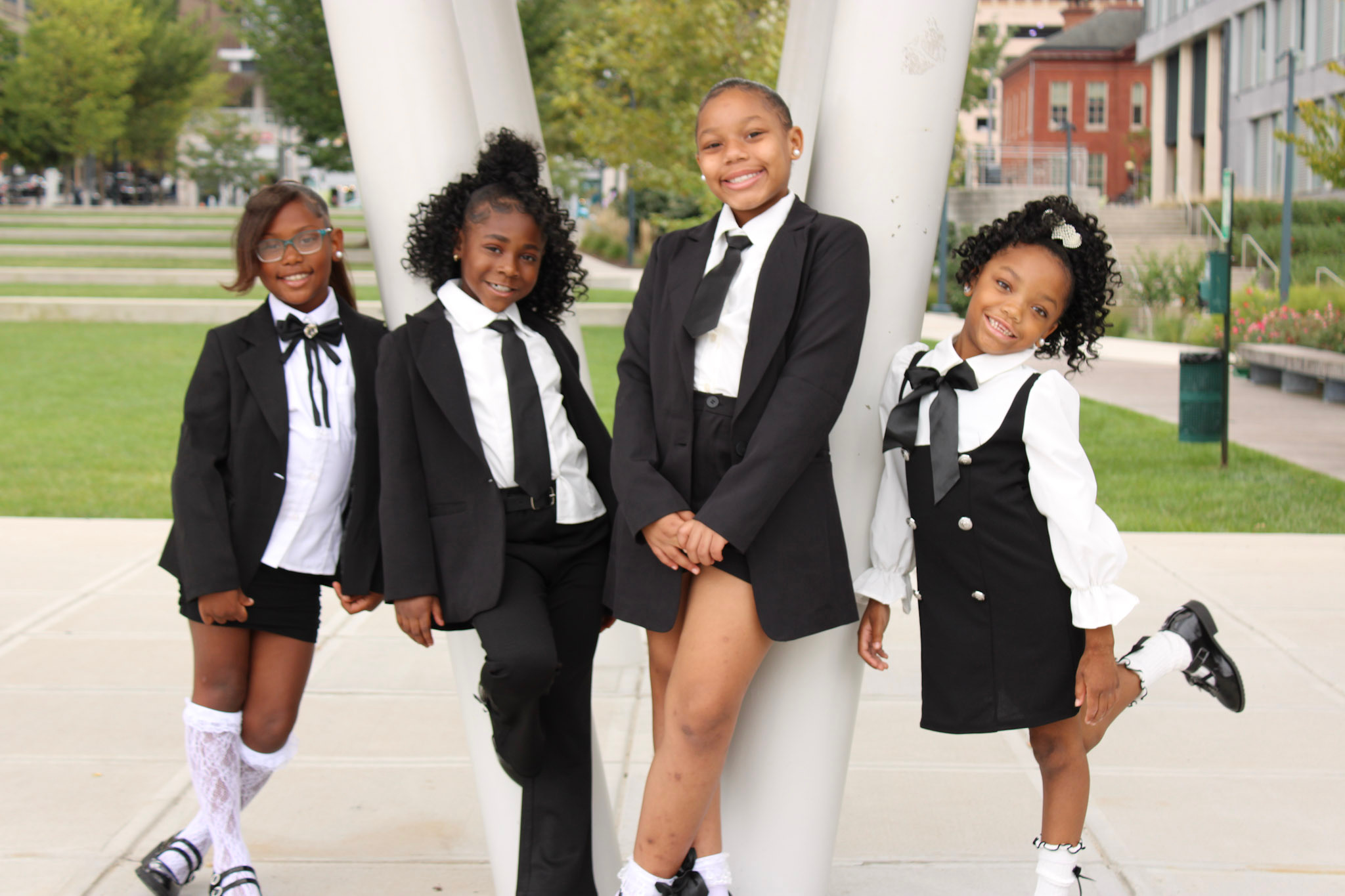 Four children in school uniforms standing outdoors on a paved area with grass and buildings in the background.