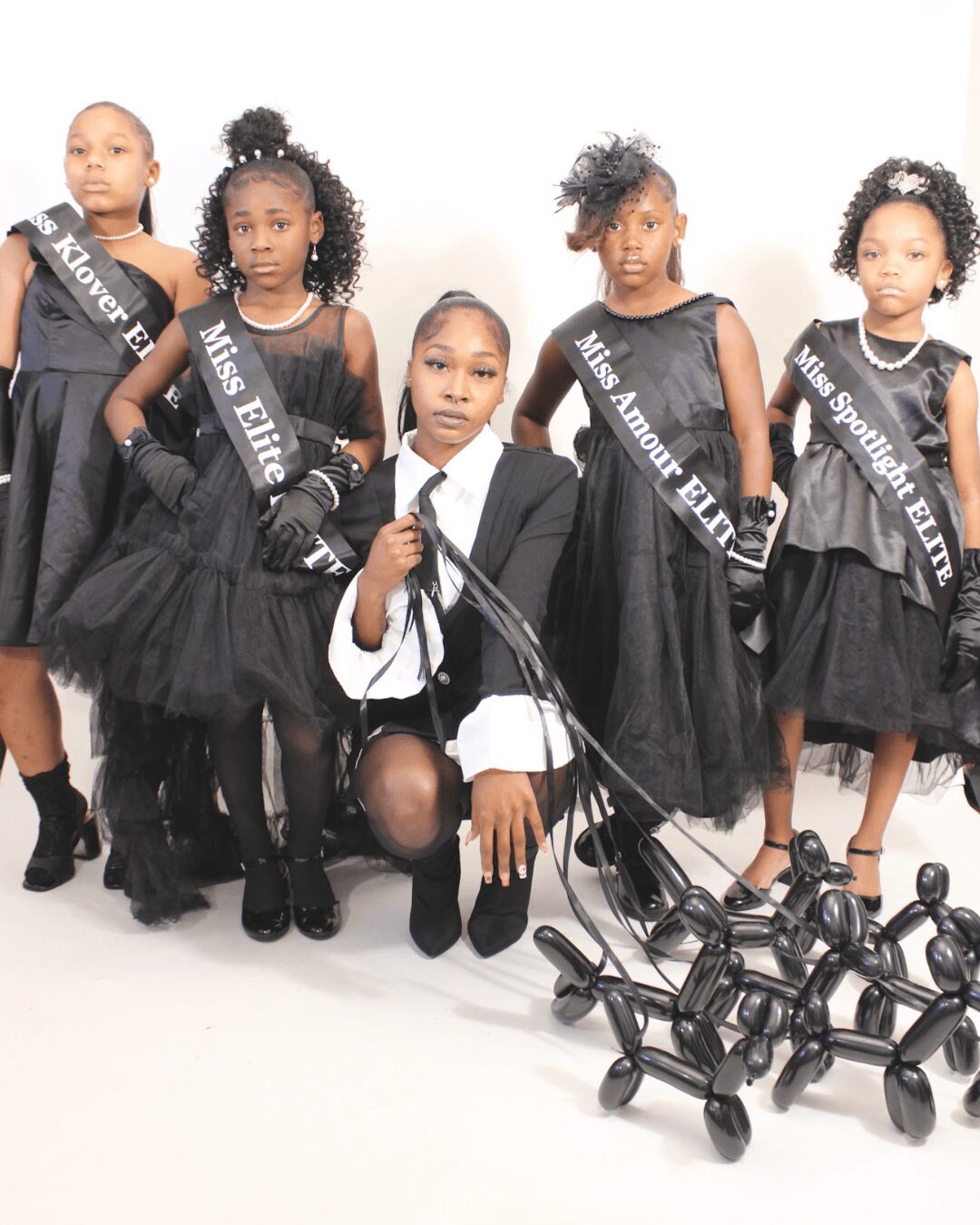 Five young girls in black dresses and sashes, with one girl kneeling and holding a cane, against a white background.