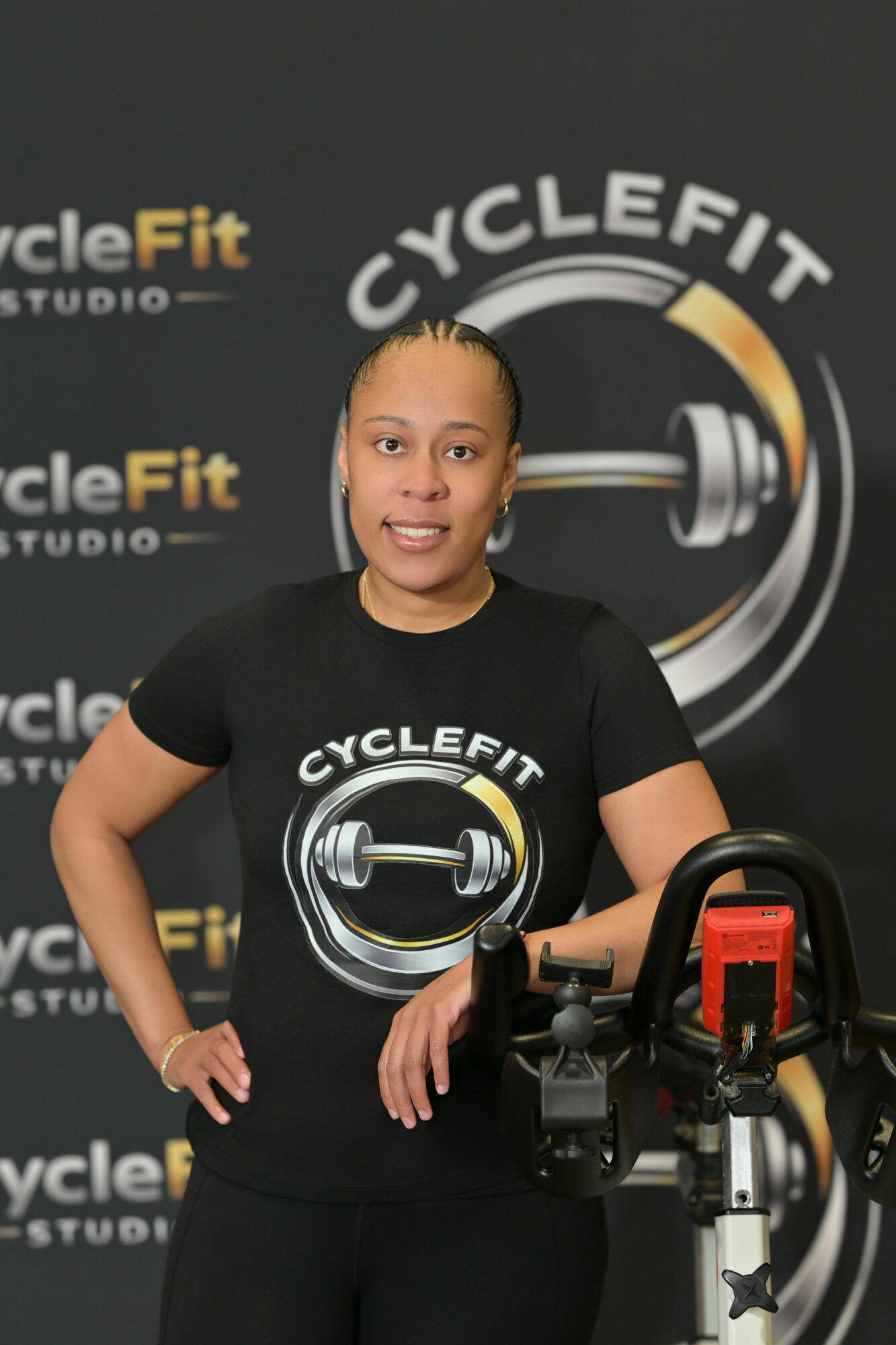 Woman standing in front of a logo background, wearing a black T-shirt with a logo, smiling, with fitness equipment.