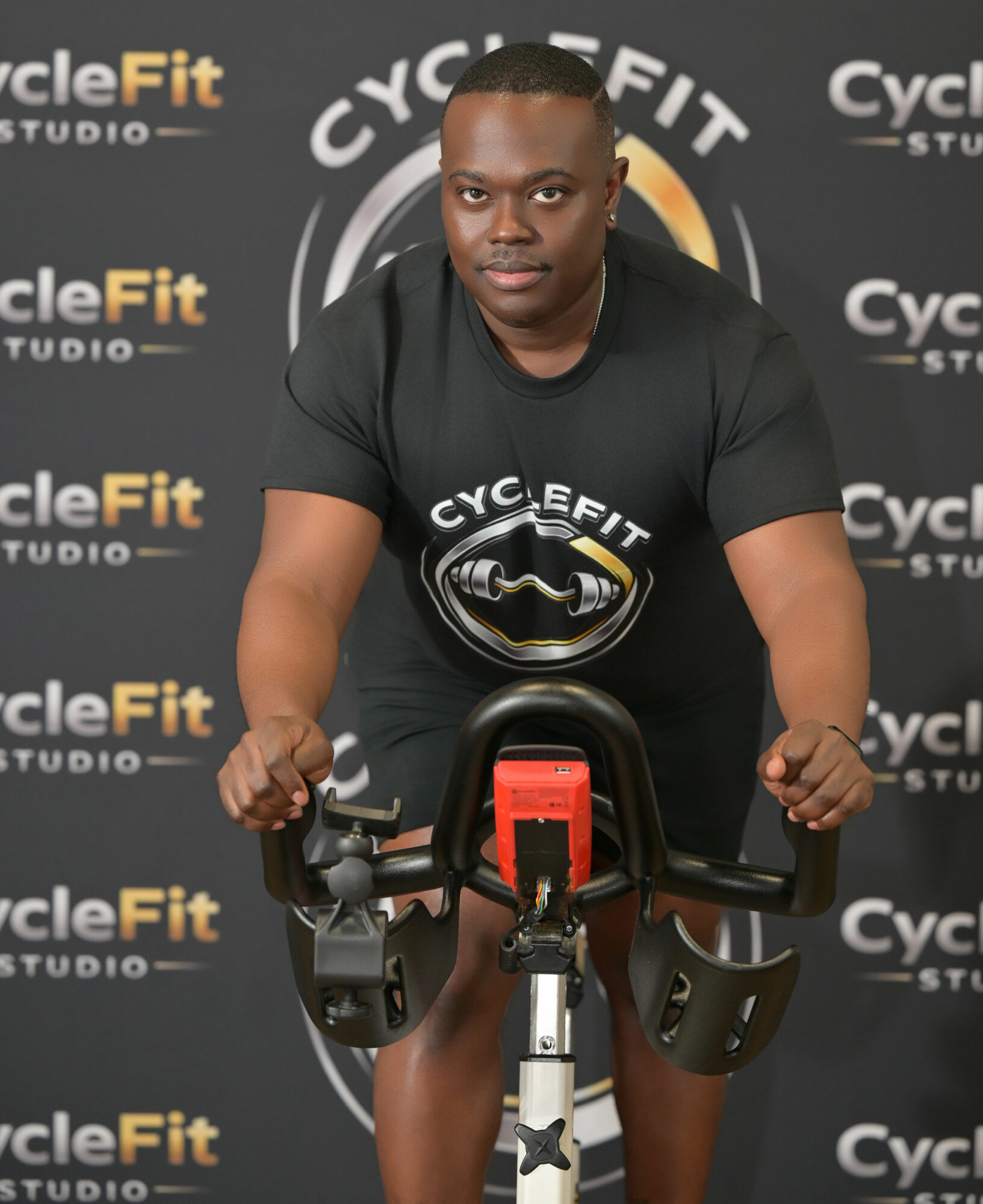 Man on exercise bike in gym with CycleFit Studio background, wearing black shirt, looking at camera.