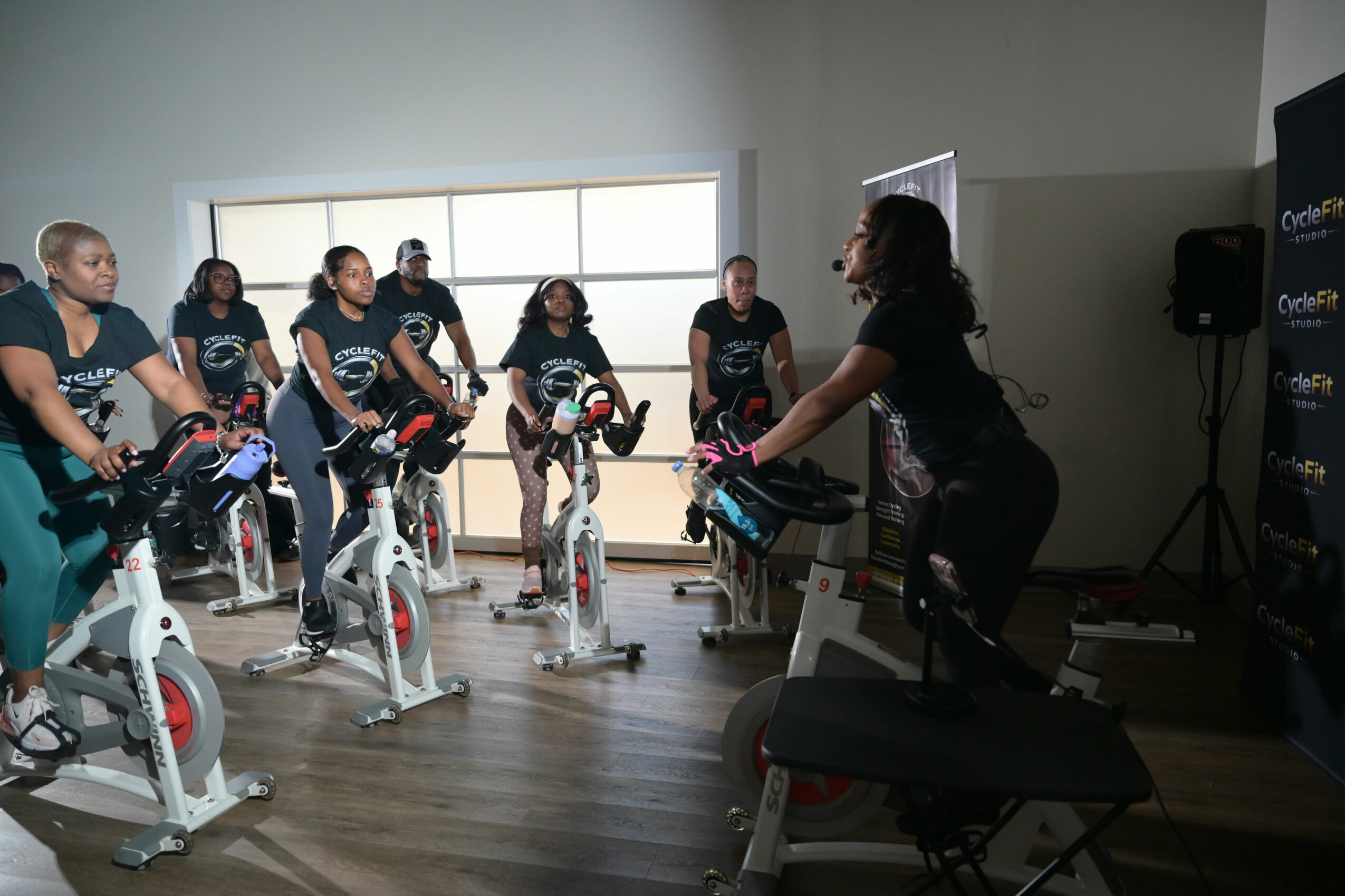 Group of people on stationary bikes listening to instructor in indoor room with large window.
