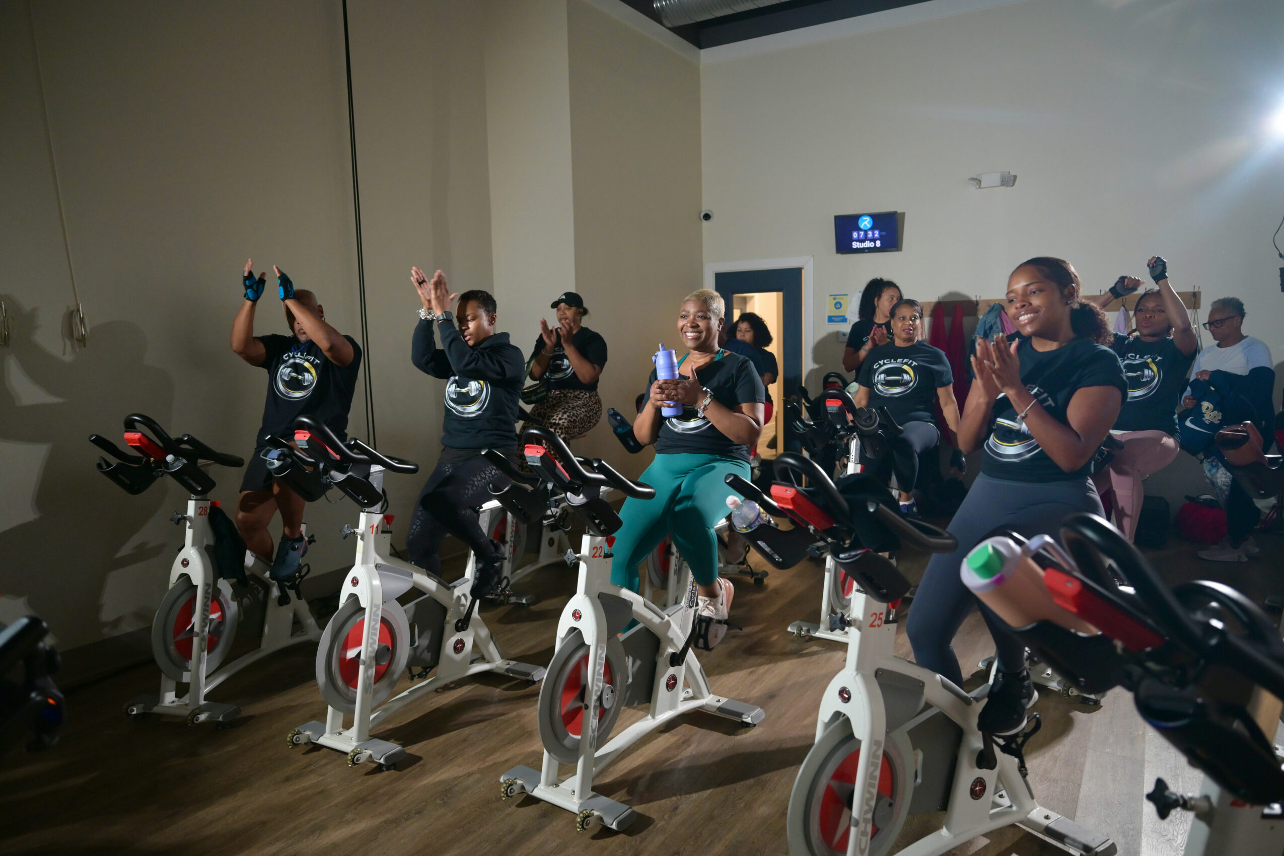 Group of people participating in indoor cycling class, some raising hands, others pedaling on stationary bikes, in a fitness studio.