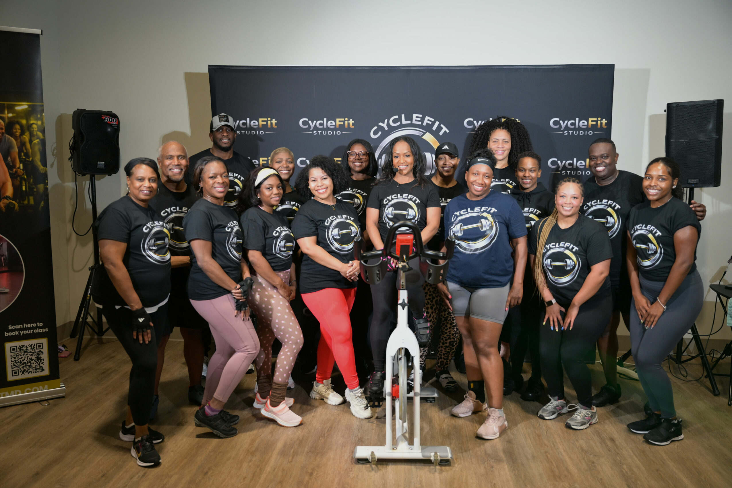 Group of people in matching shirts posing indoors with cycling equipment and a backdrop, smiling for the photo.