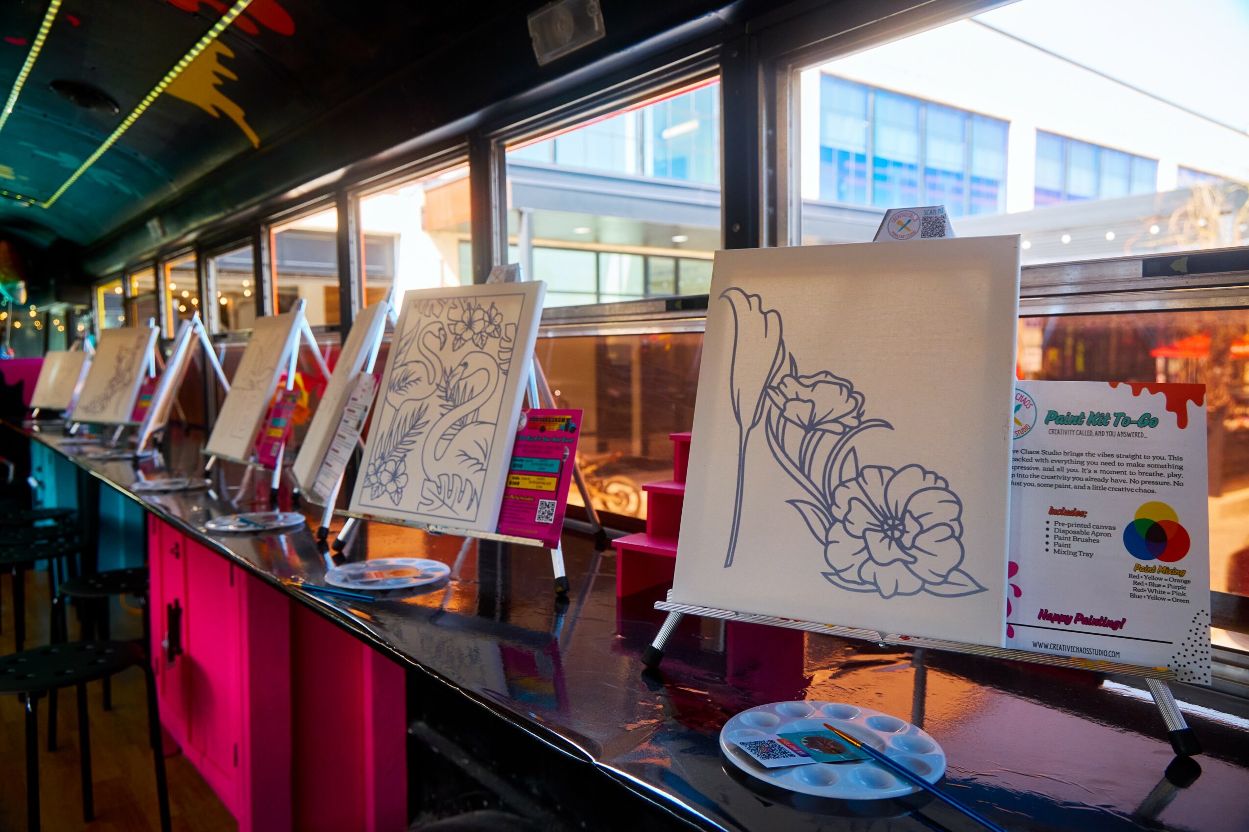 Art display with sketches and paintings on a long table inside a building with large windows.