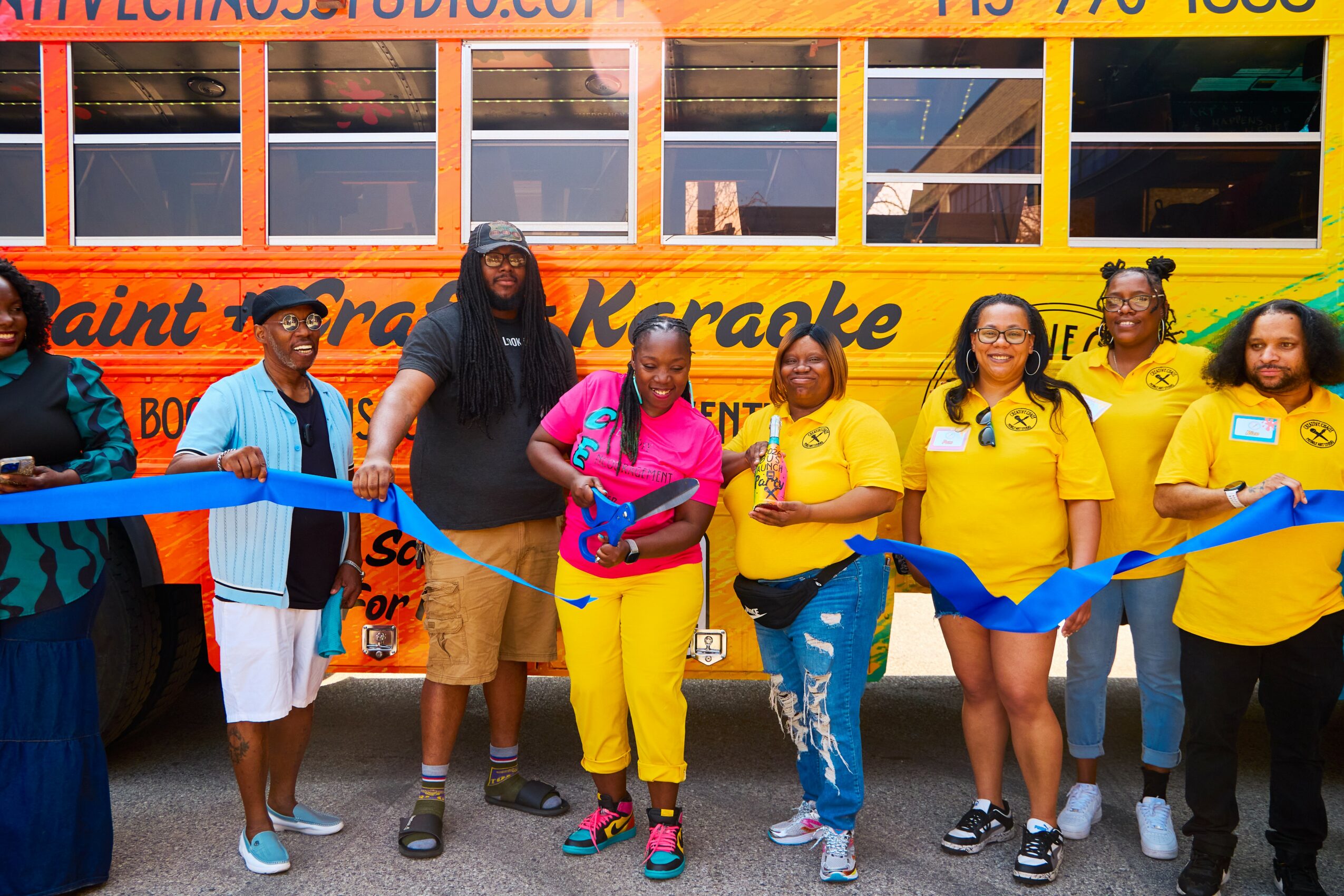 Group of people standing in front of a colorful bus, holding a blue ribbon for a ribbon-cutting event.