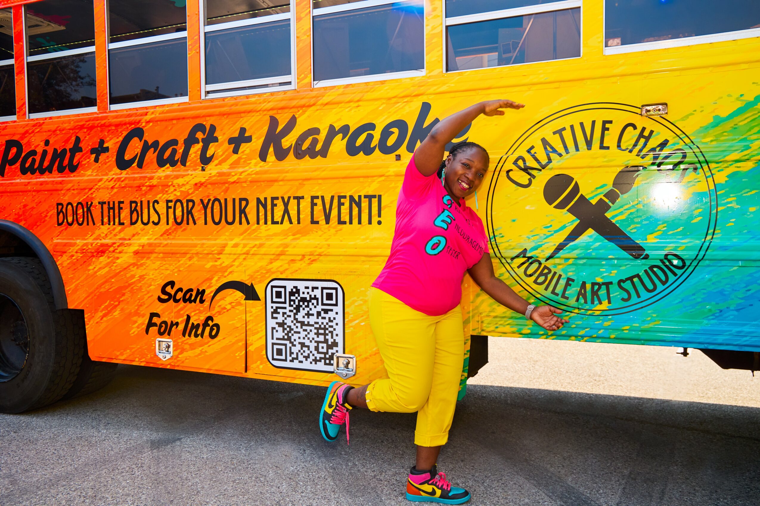 Person in pink shirt and yellow pants poses next to colorful bus with creative art studio logo.