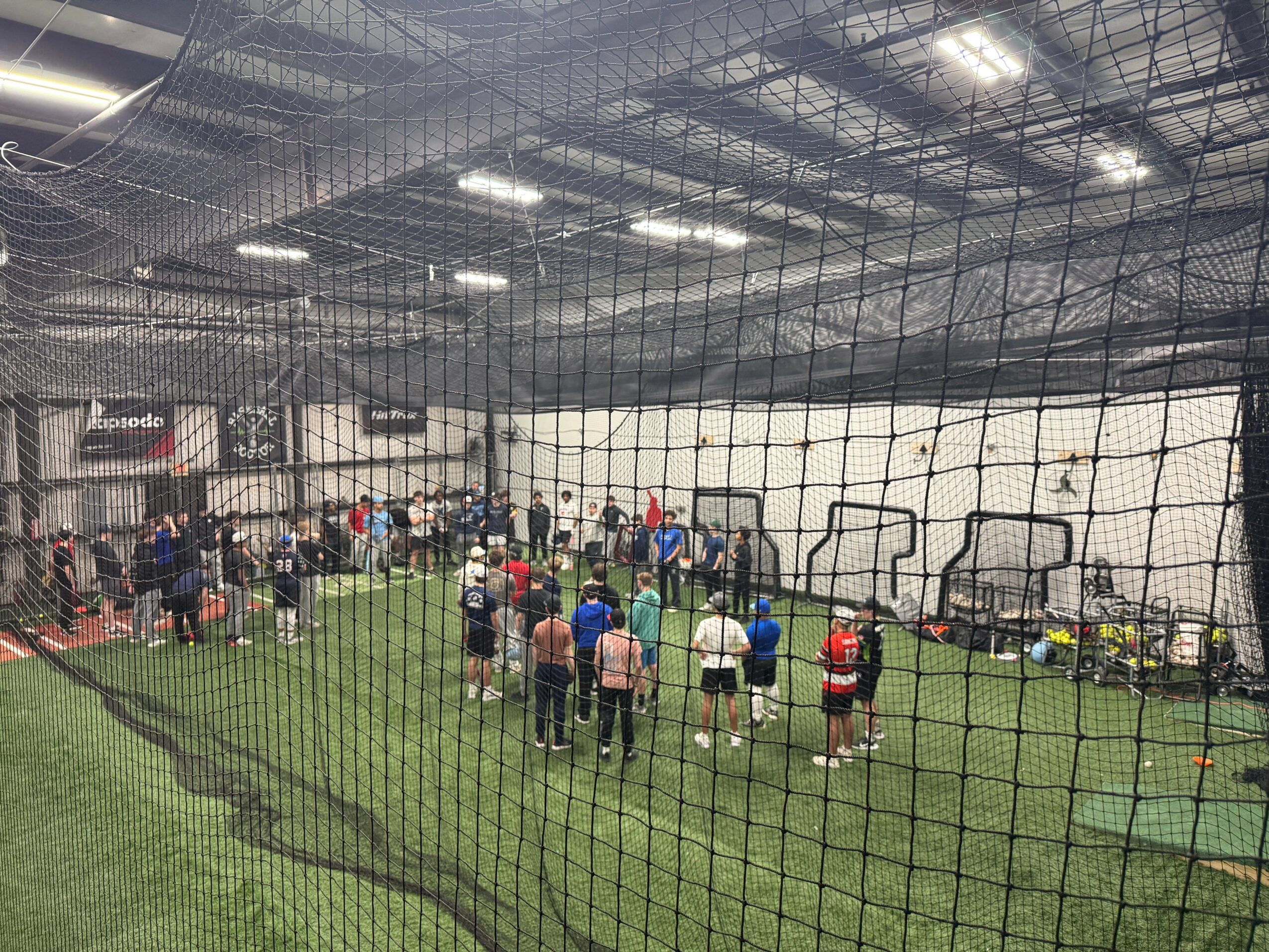Indoor sports facility with people standing on artificial turf, surrounded by a netted barrier.
