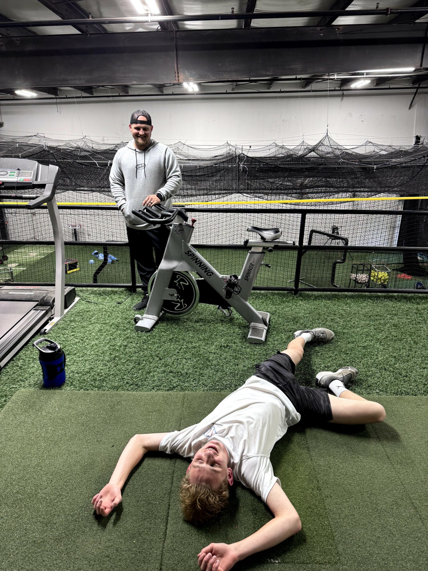 Person lying on the ground with a person standing behind a stationary exercise bike in an indoor gym.