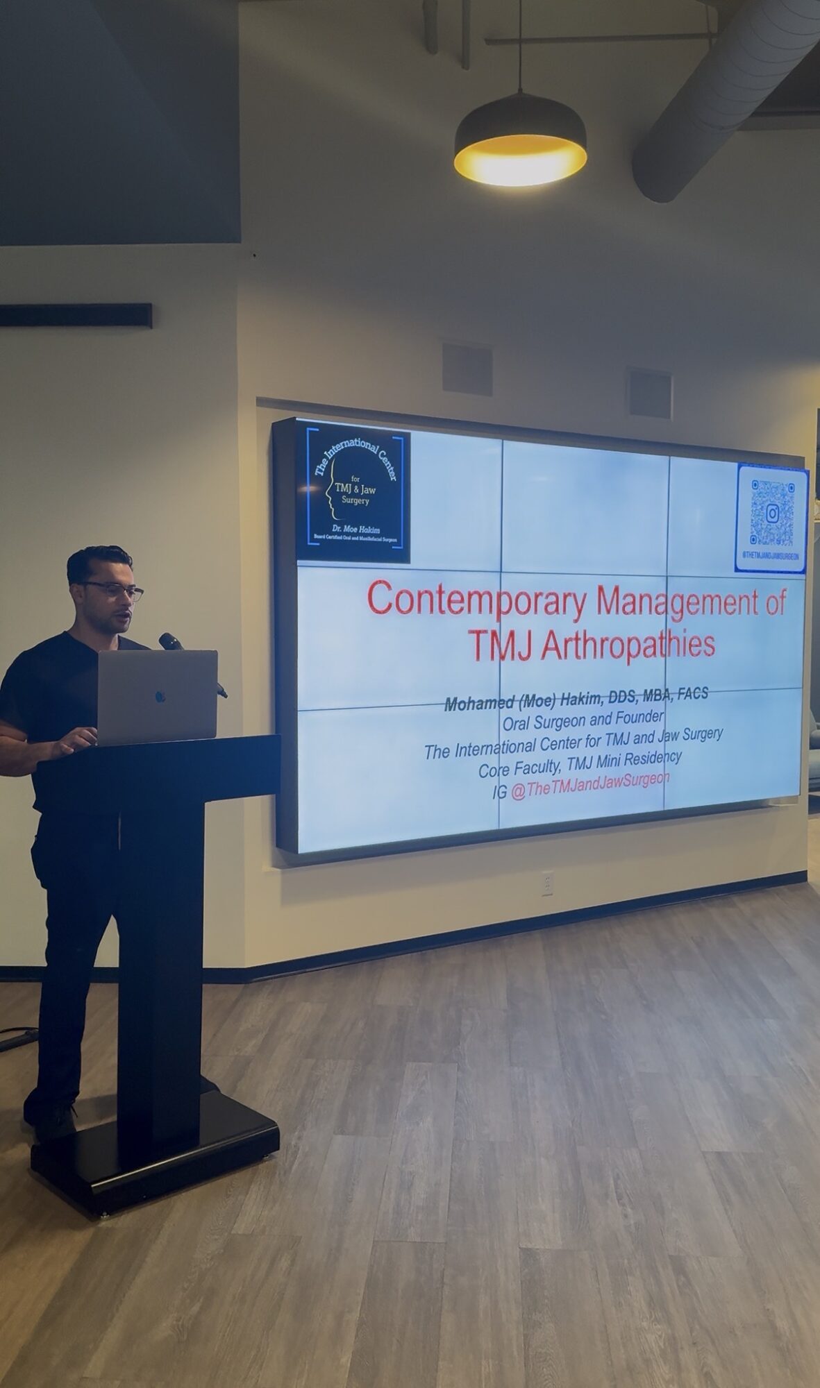 Person standing at podium giving a presentation in a room with a large screen displaying a slide titled 'Contemporary Management in TMJ Arthroplasty'.