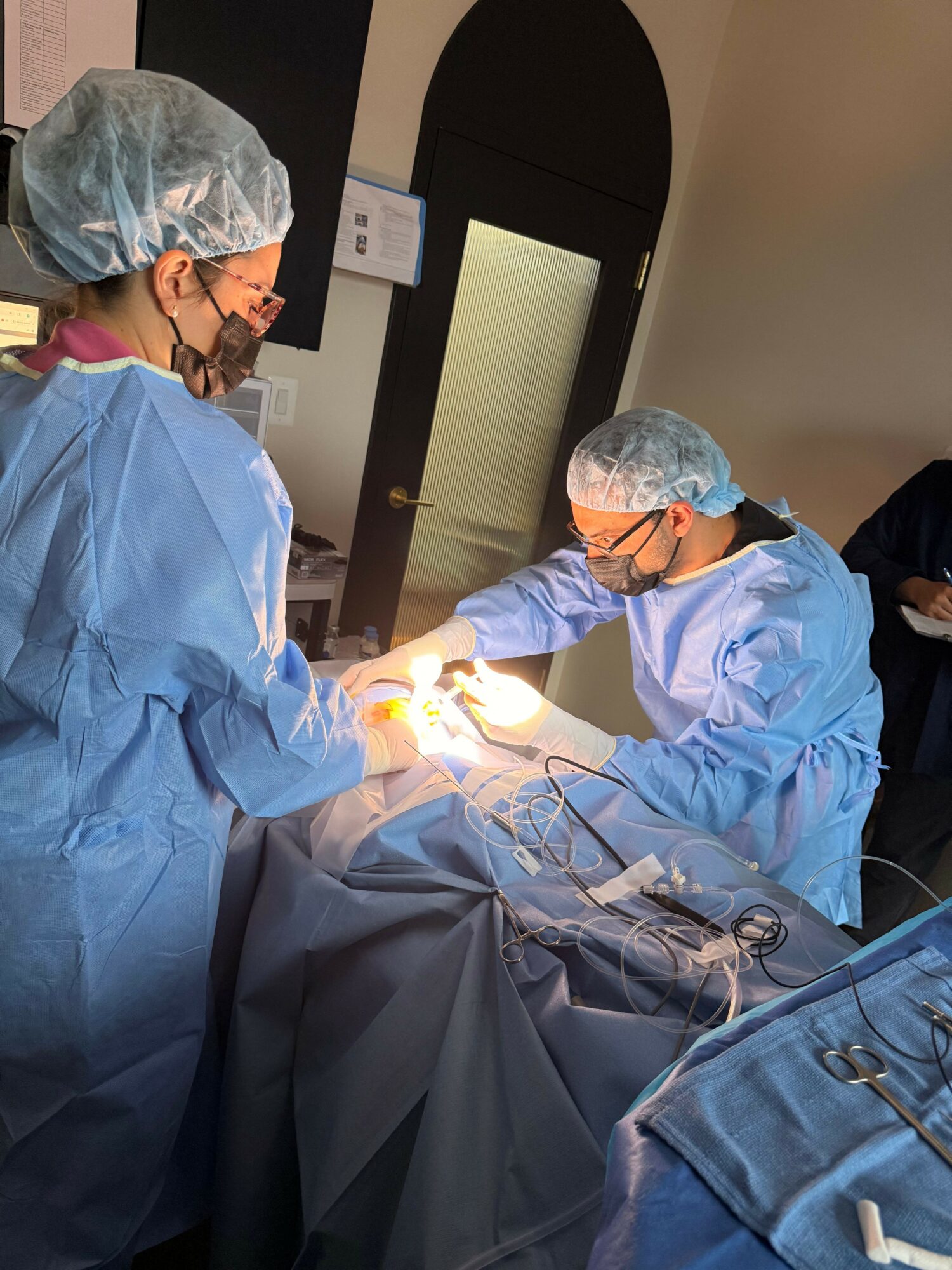 Two medical professionals in scrubs, masks, and hair covers perform surgery in an operating room.