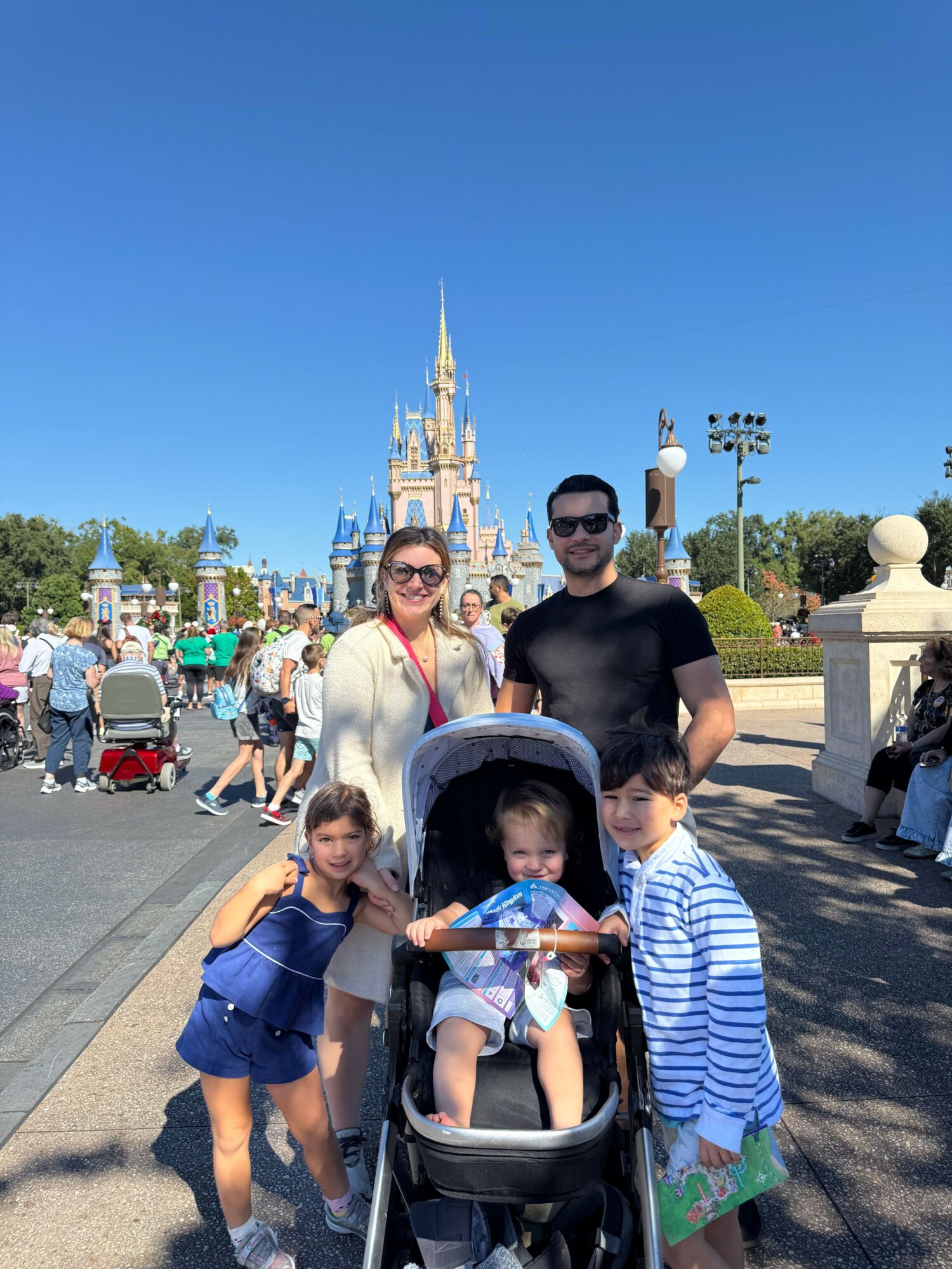Family of four with stroller in front of Disney castle, sunny day, people walking around.