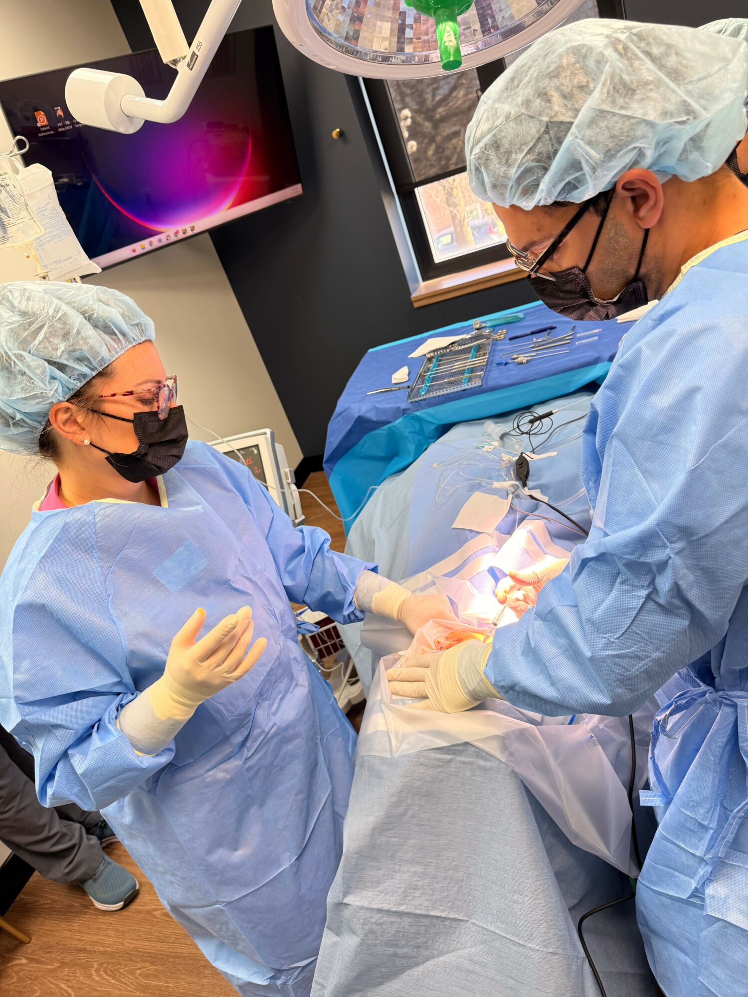 Two medical professionals in surgical attire perform a procedure in an operating room with monitors and equipment.