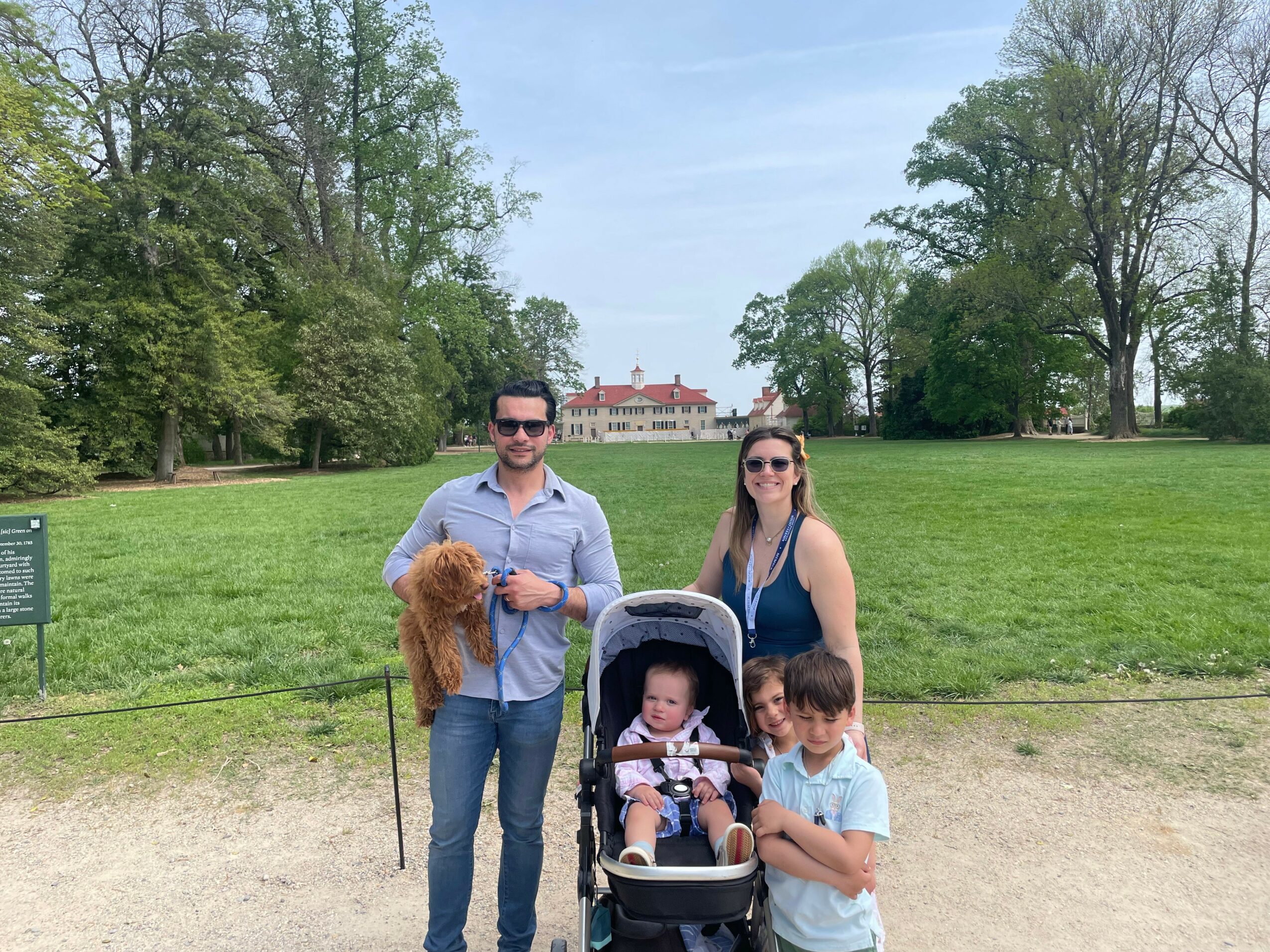Family with two children standing on a grassy park with trees and a building in the background.