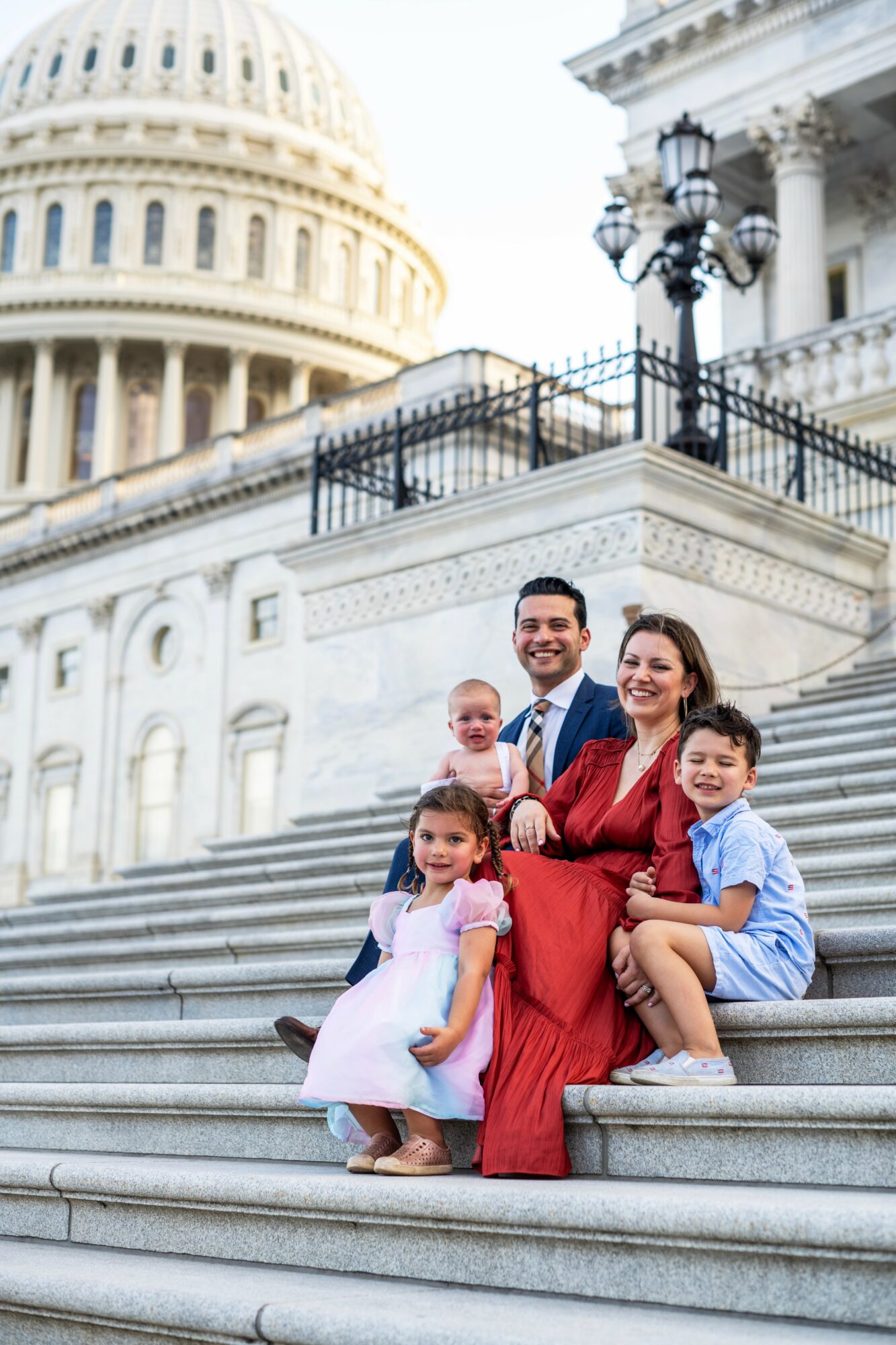Family of five sitting on steps outside government building, smiling, children with casual clothes, adults in formal attire.