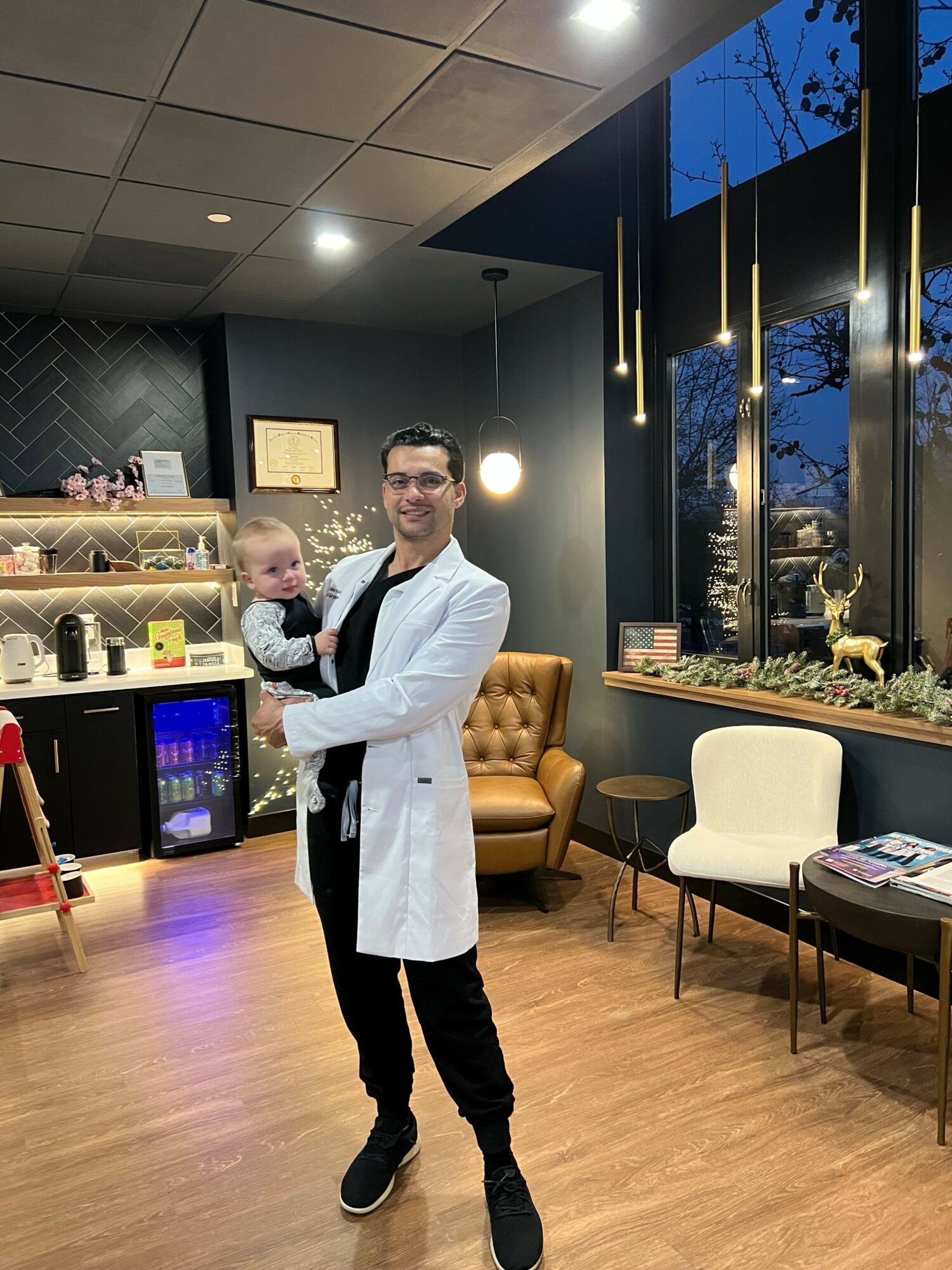 Man in white coat holding a baby in a modern indoor space with chairs and large windows.
