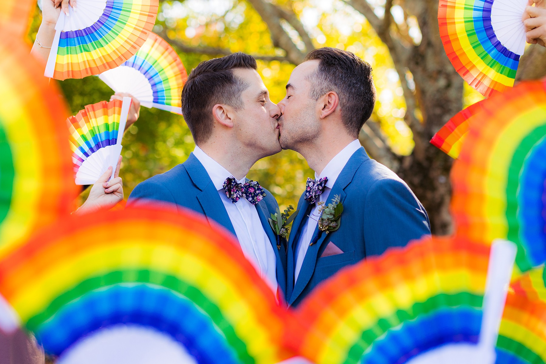 Two men in suits kiss surrounded by rainbow-colored fans outdoors with trees in the background.