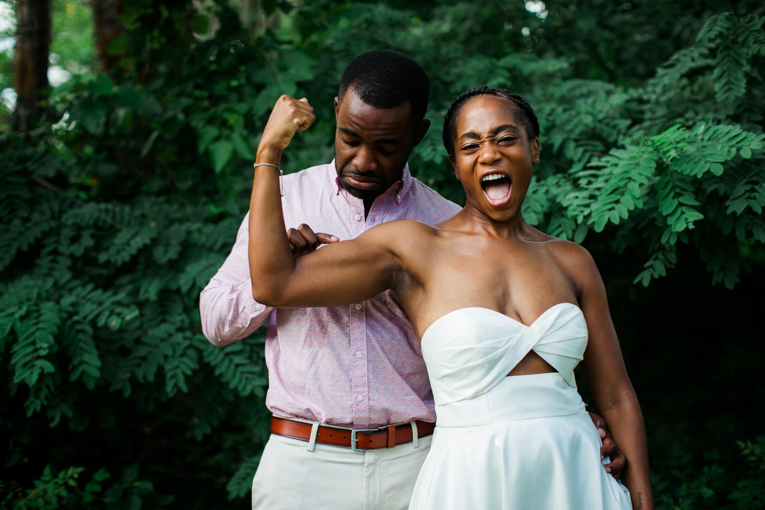 Two people outdoors, one flexing arm and the other smiling widely, surrounded by green foliage.