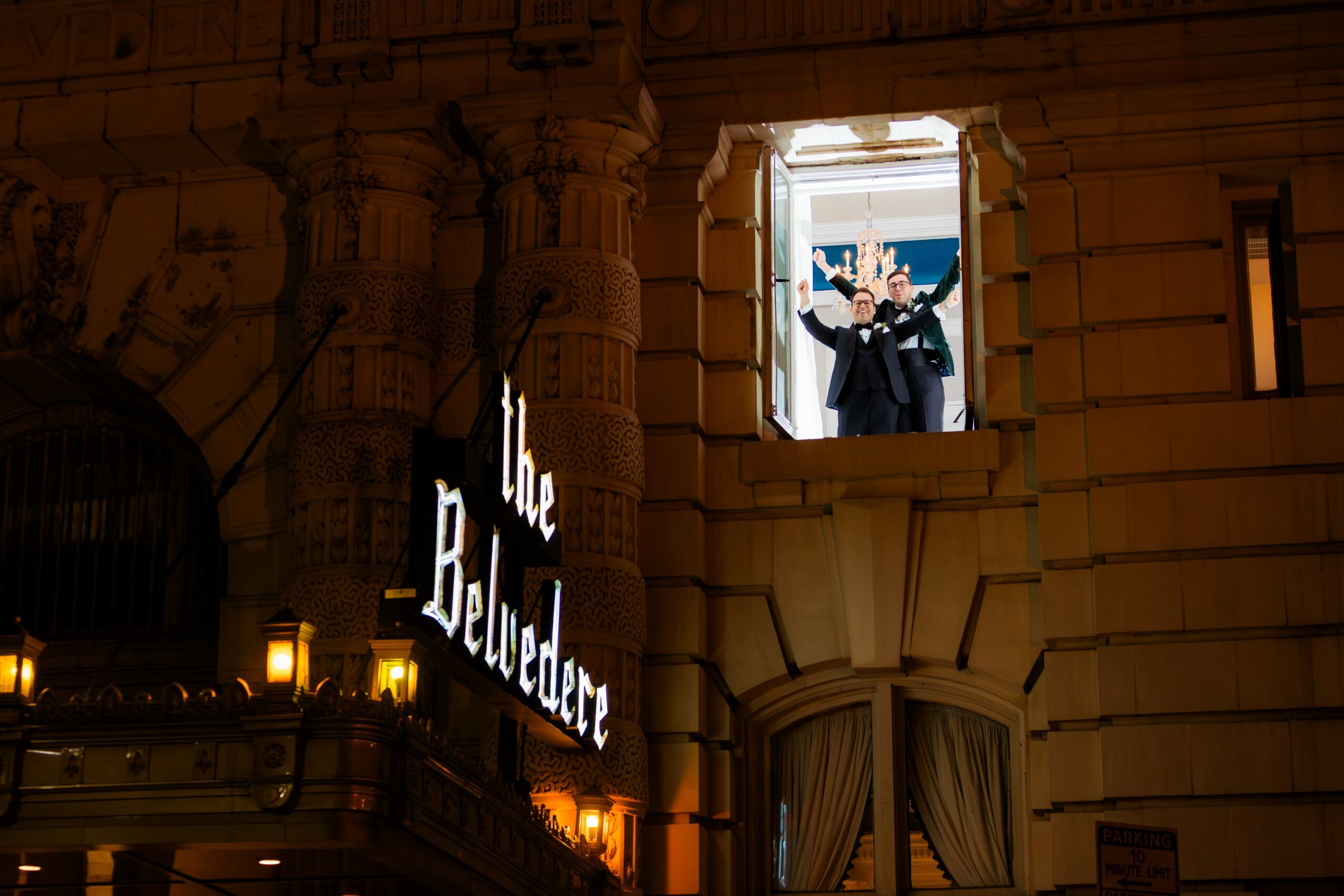Person standing at a window on a building's upper floor, holding a camera or phone, with a cityscape background.