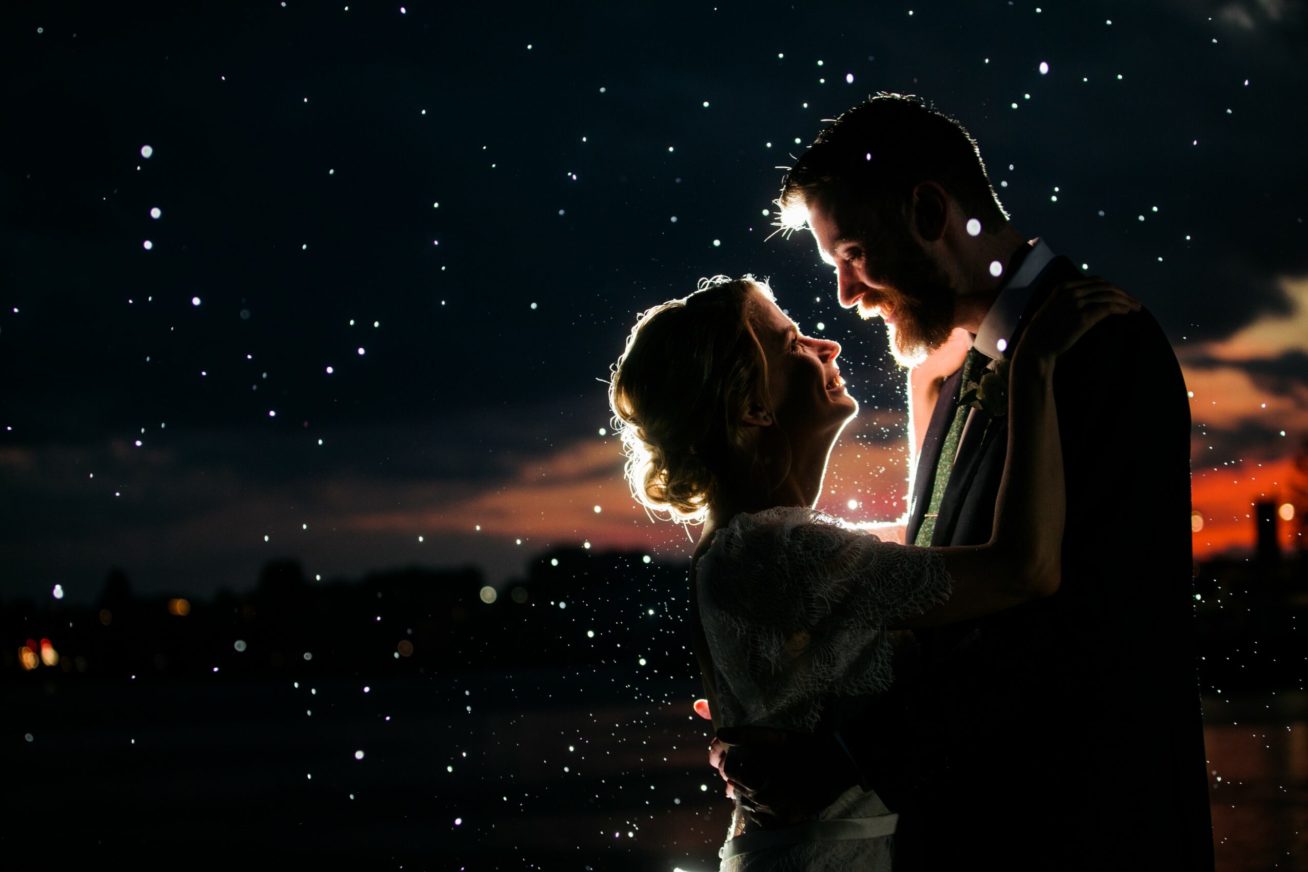 A couple stands close together outdoors at night with stars in the sky, backlit by a sunset or city lights.