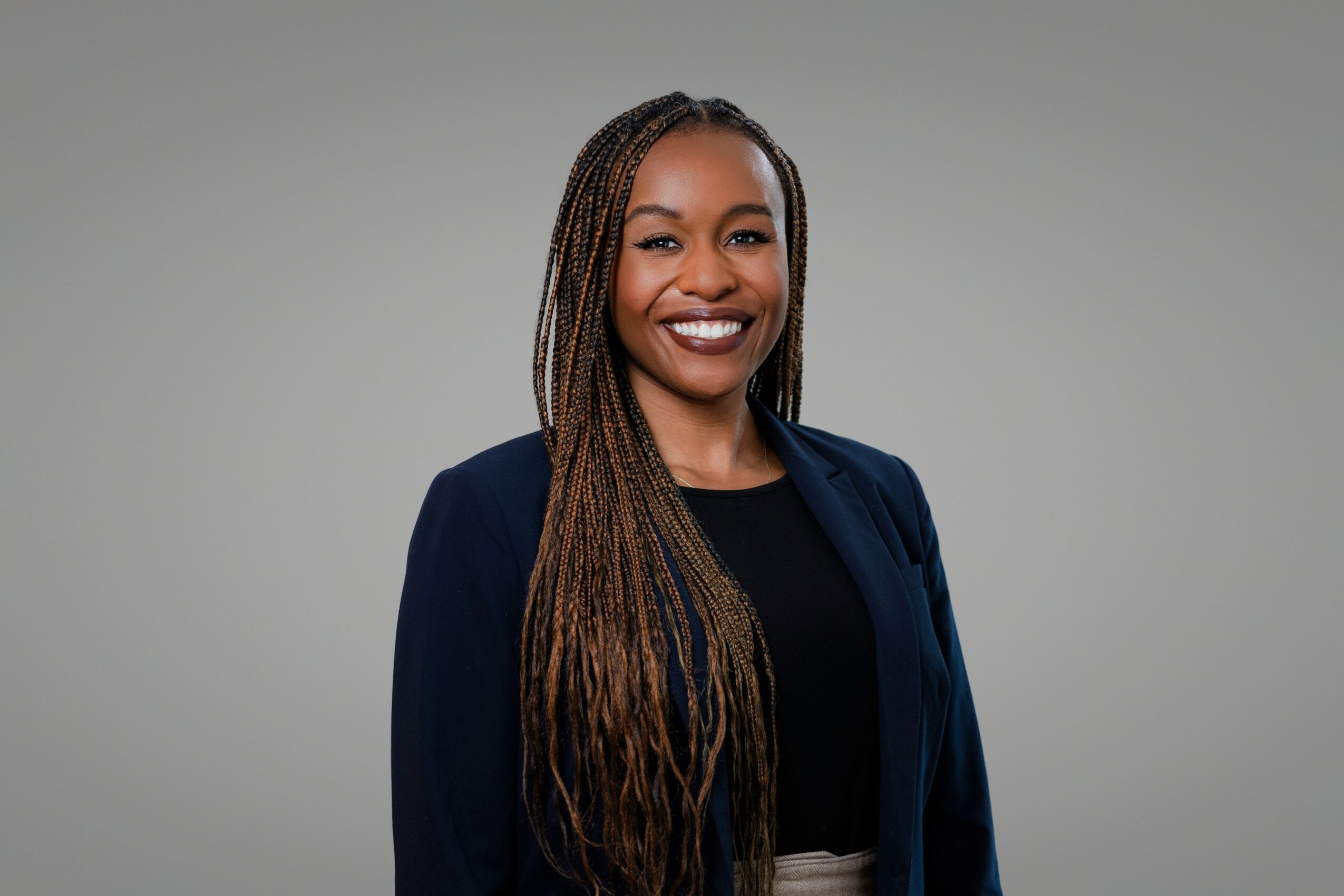 Smiling woman with long braided hair wearing a dark blazer and black top, standing against a gray background.