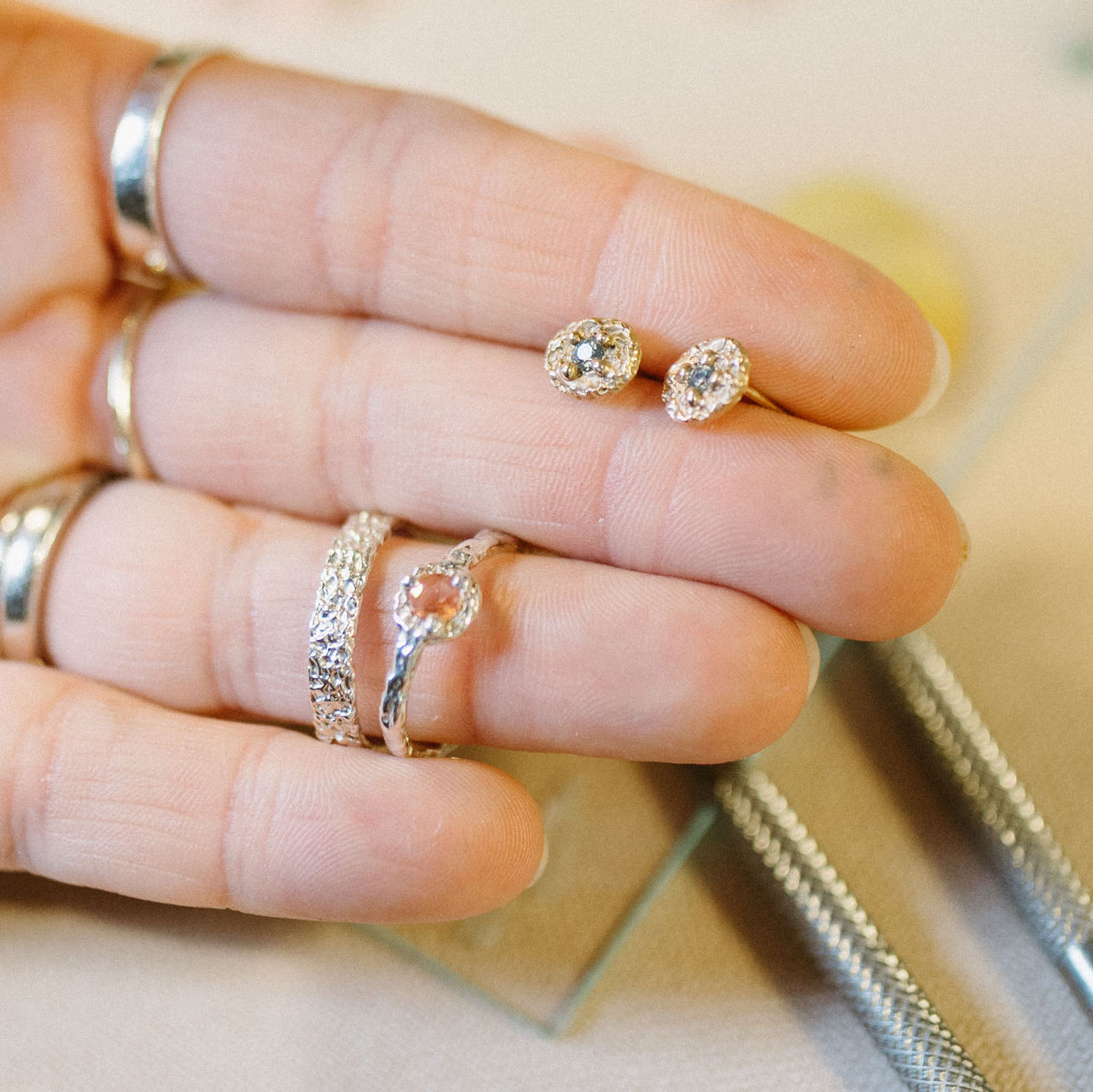 Hand with multiple rings, including diamond-studded and plain bands, resting on a surface with jewelry tools nearby.