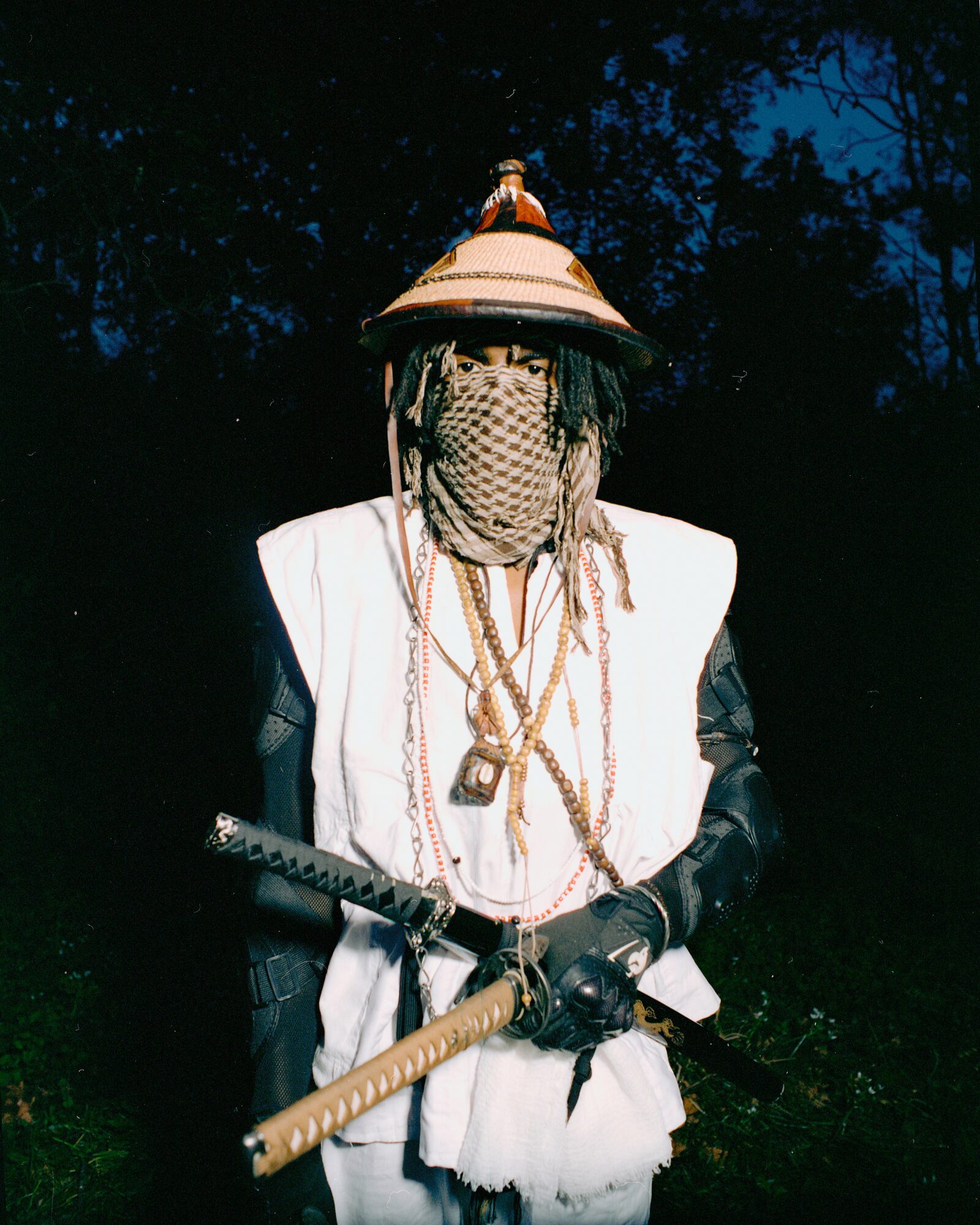 Person wearing a hat, face covered with a cloth, holding a sword, outdoors at night.