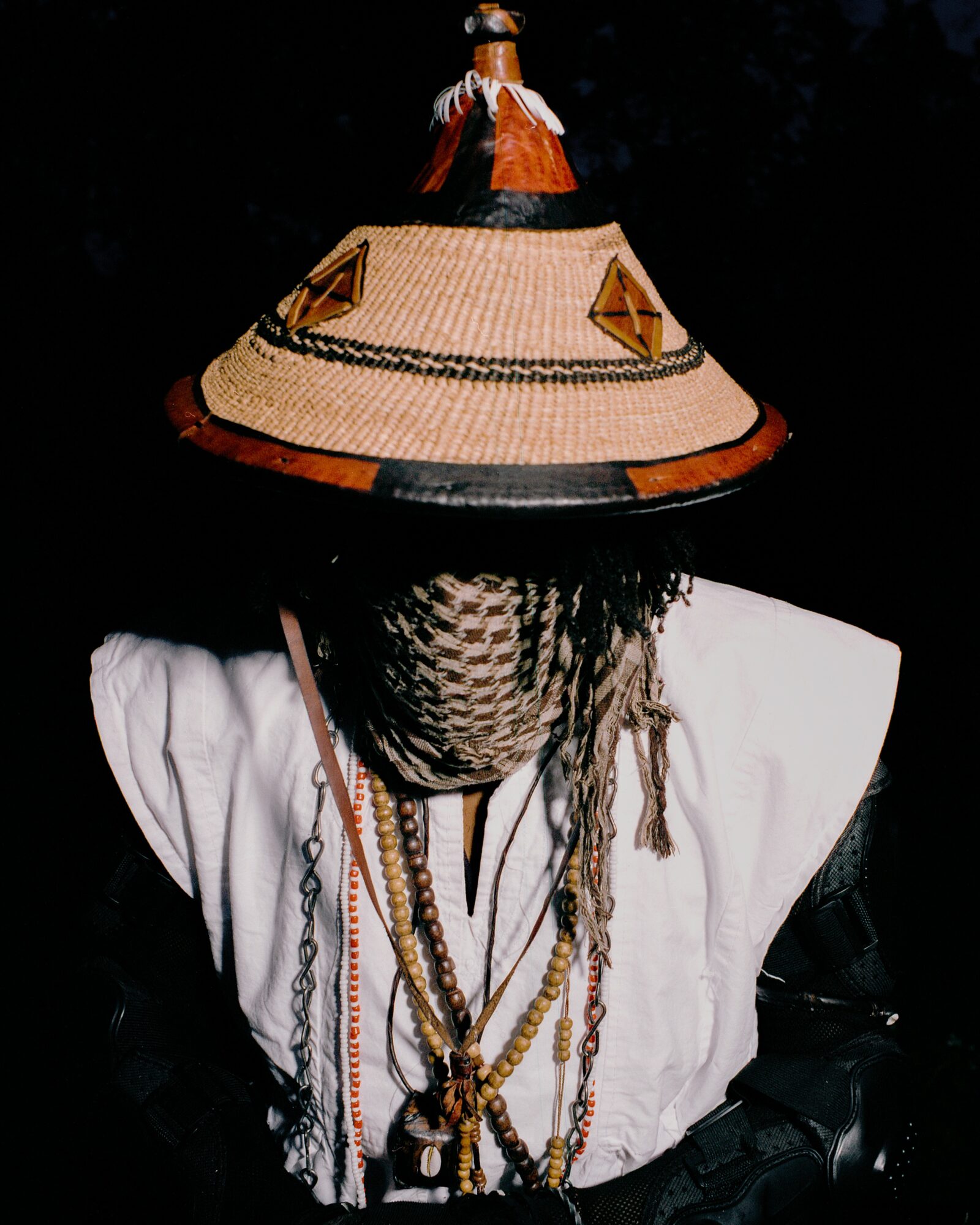 Person wearing a large, decorated hat with a figure on top, and multiple necklaces, dressed in white and black clothing.