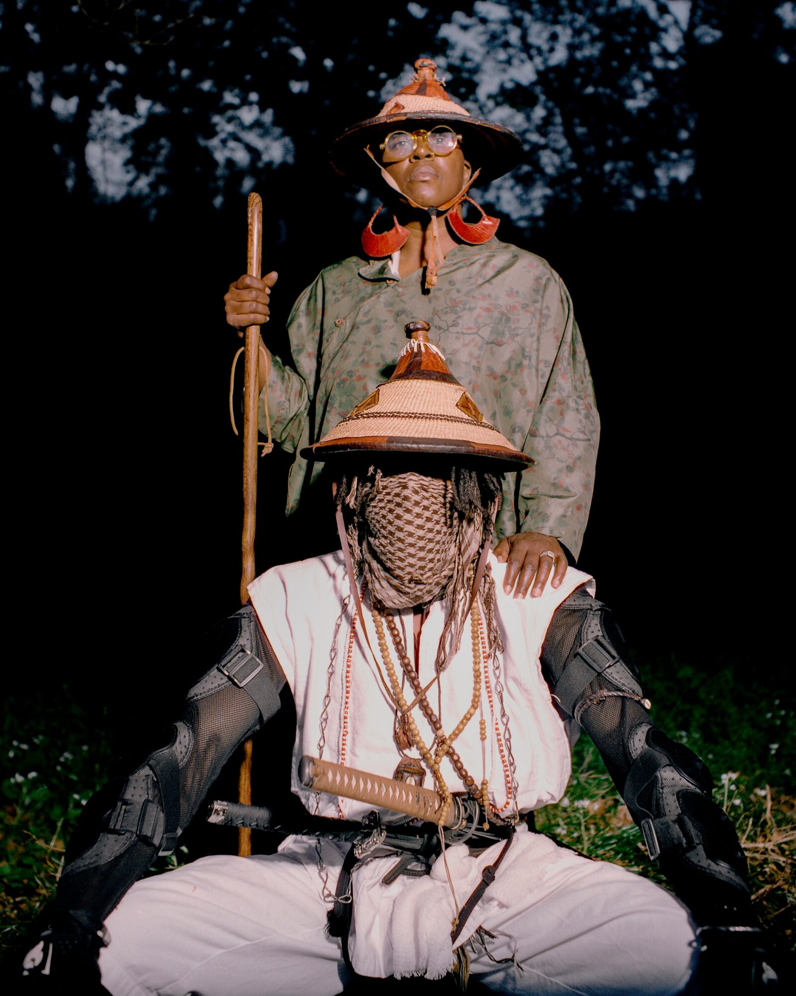 Two individuals in traditional attire outdoors, one standing with a staff, the other sitting with head covered and jewelry.