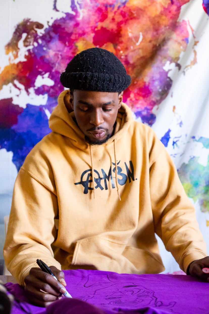Young man in yellow hoodie and black beanie writing on purple fabric, colorful abstract background behind him.