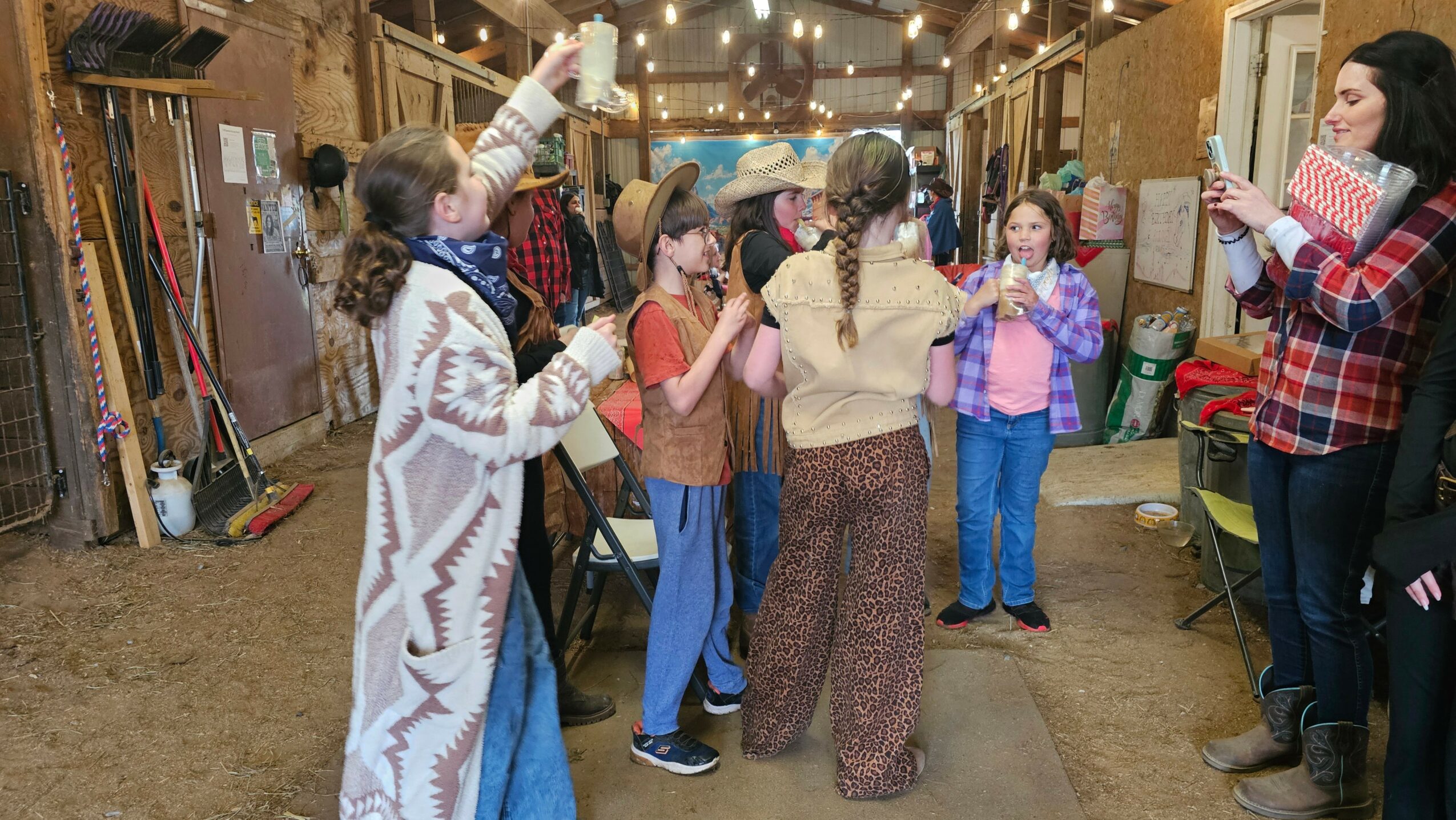 Group of children and adults in a barn-like setting, some taking photos, others talking and interacting.