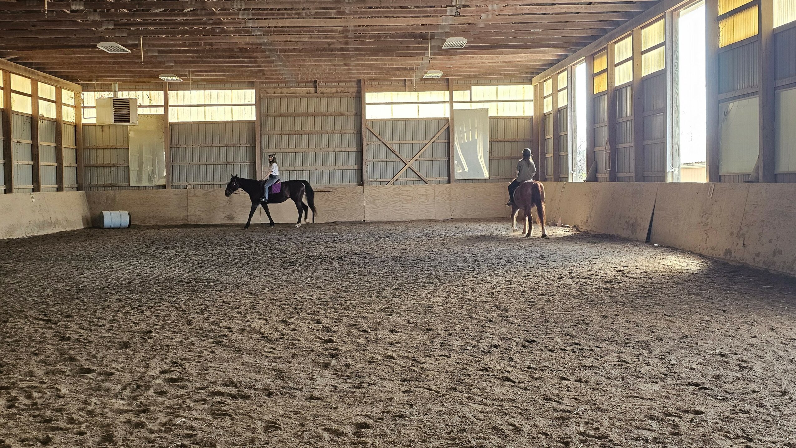 Two people riding horses inside an indoor riding arena with high windows and a dirt floor.