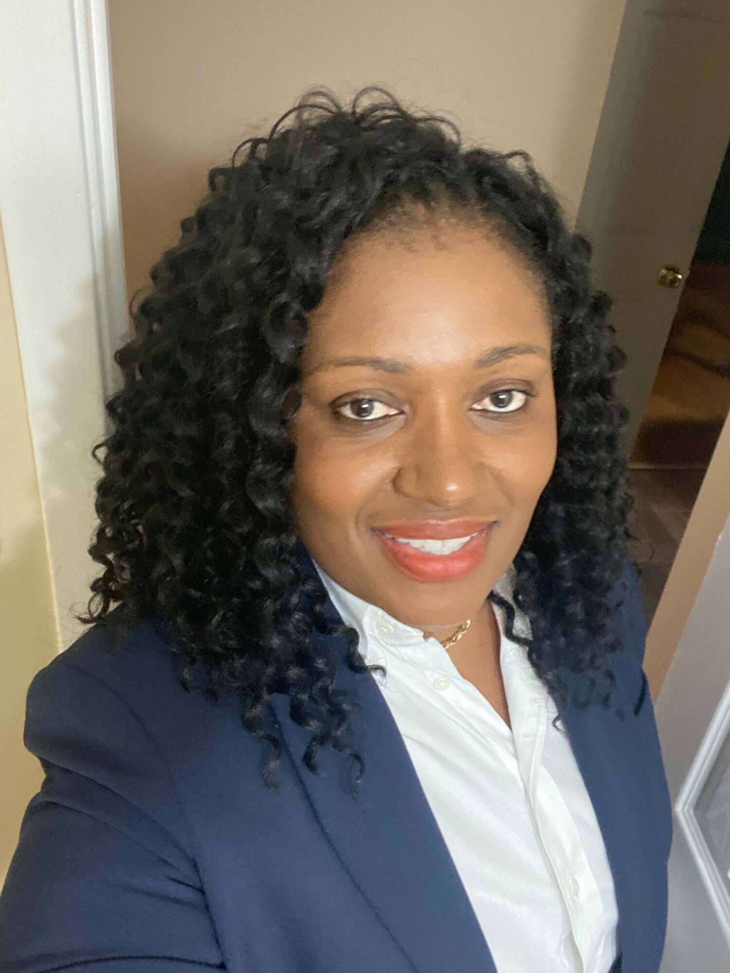 Woman with curly hair wearing a white shirt and navy blazer, smiling indoors.