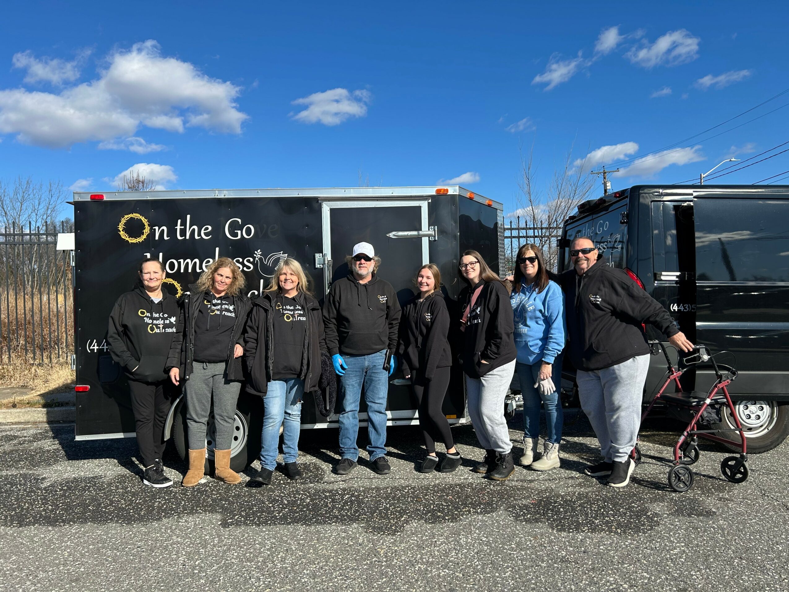 Group of nine people standing outdoors in front of a black trailer, with a blue sky and clouds above.