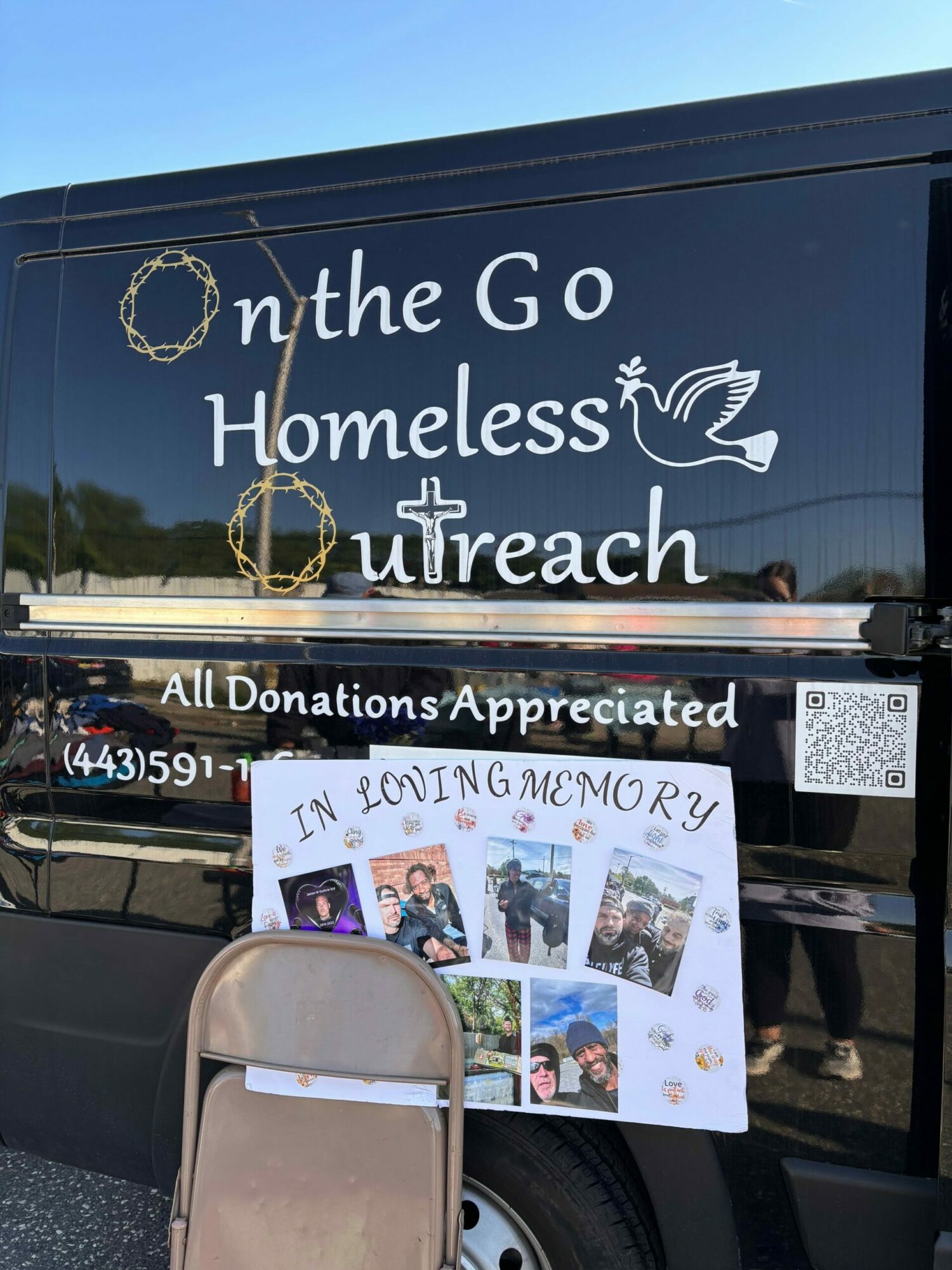 A black vehicle with white text reading 'On the Go Homeless Outreach' and a dove logo, with a donation sign and photos.