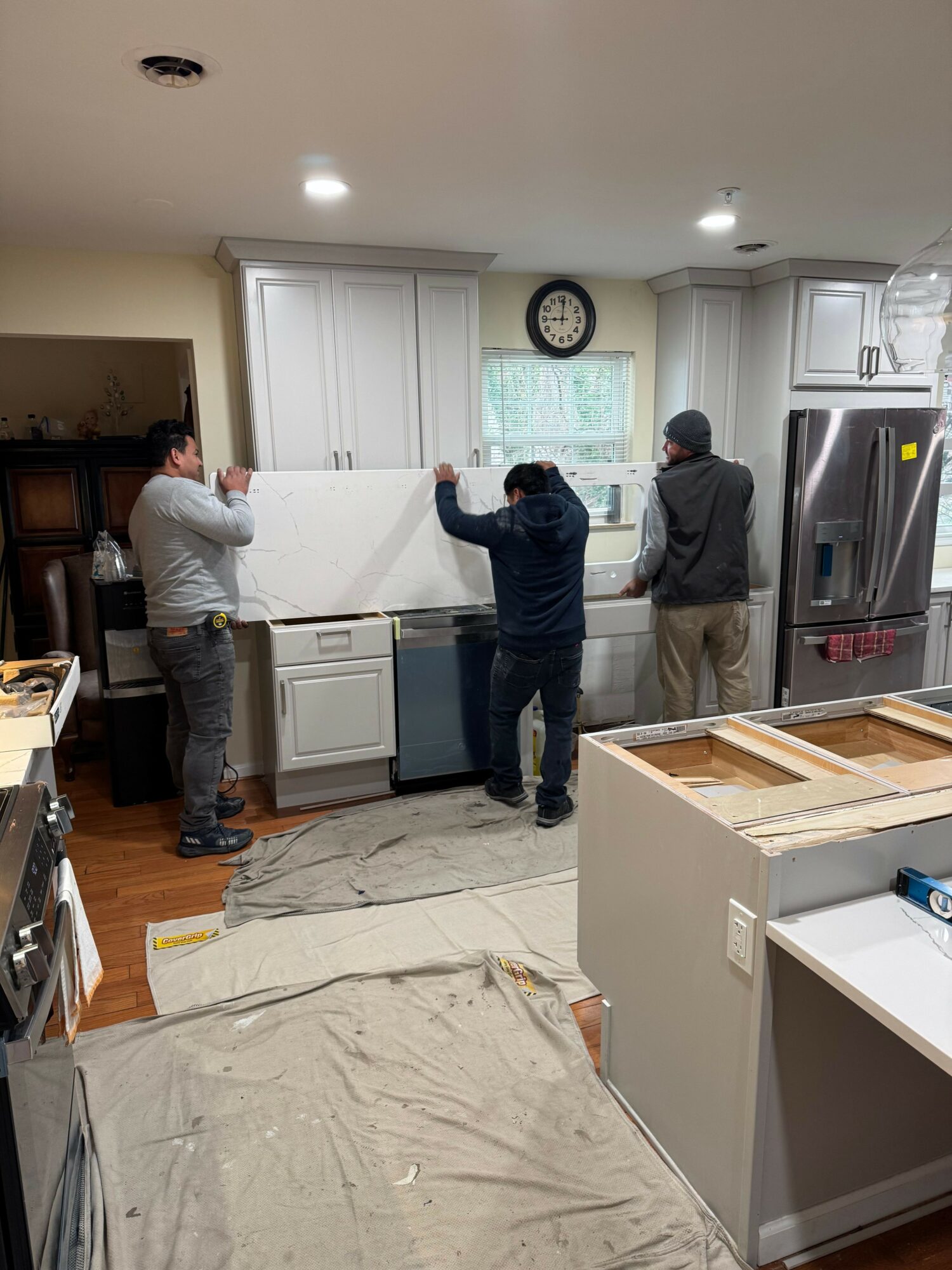 Three men working in a kitchen, installing or repairing cabinets and appliances, with a clock on the wall and a window.