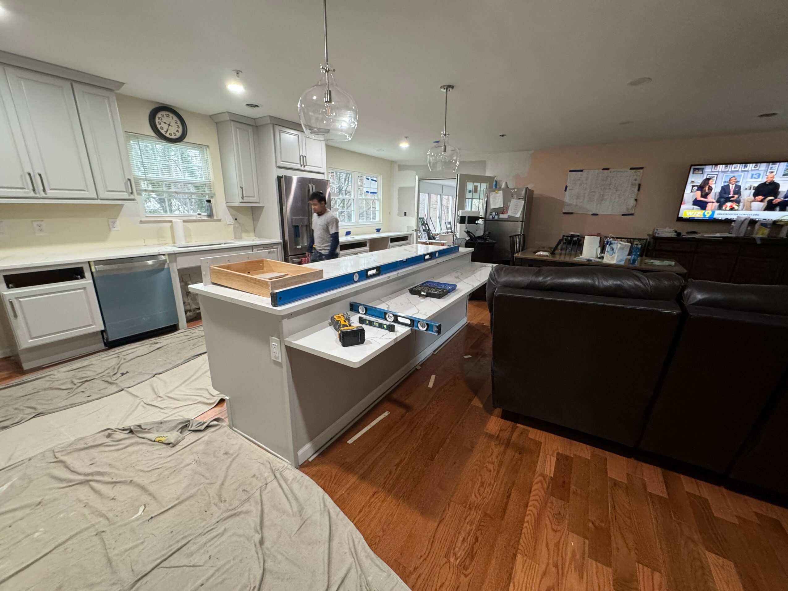 Open kitchen and living room with white cabinets, island, black sofas, and a television on the wall.