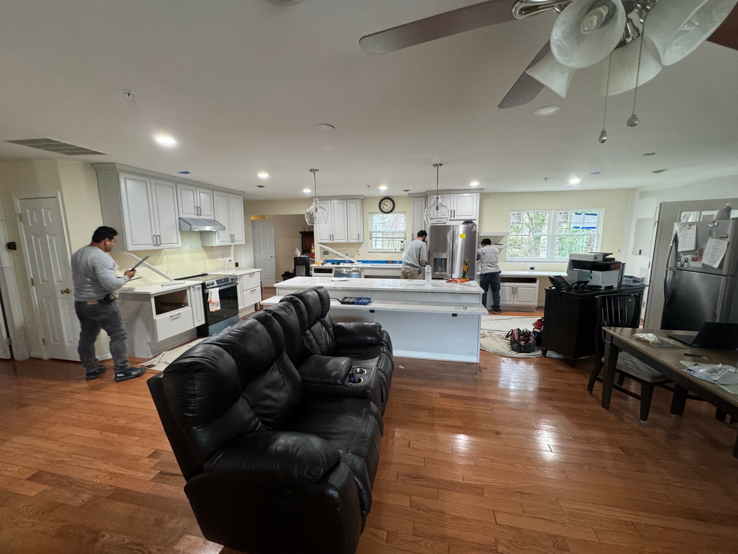 Open kitchen and living area with three people, black sofa, wooden floor, and kitchen appliances.