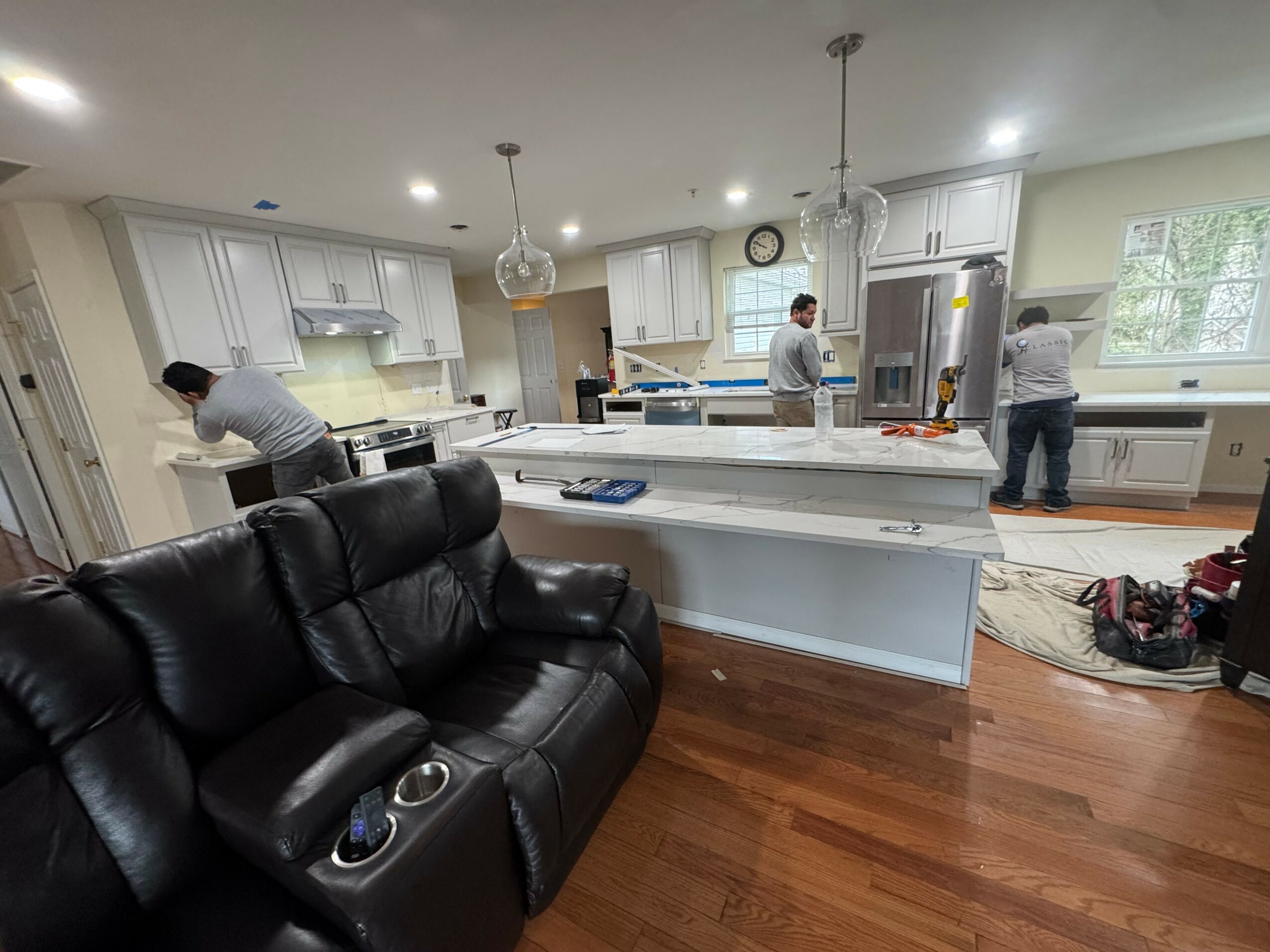 Kitchen with four people working, black sofa in foreground, white cabinets, and hardwood floor.