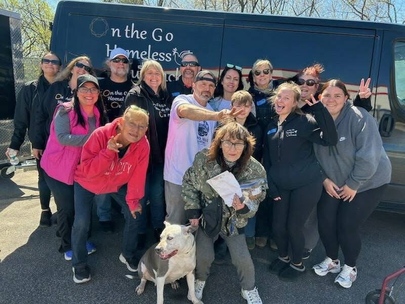 Group of people standing outdoors, some making peace signs, with a bus in the background. A dog is in front.