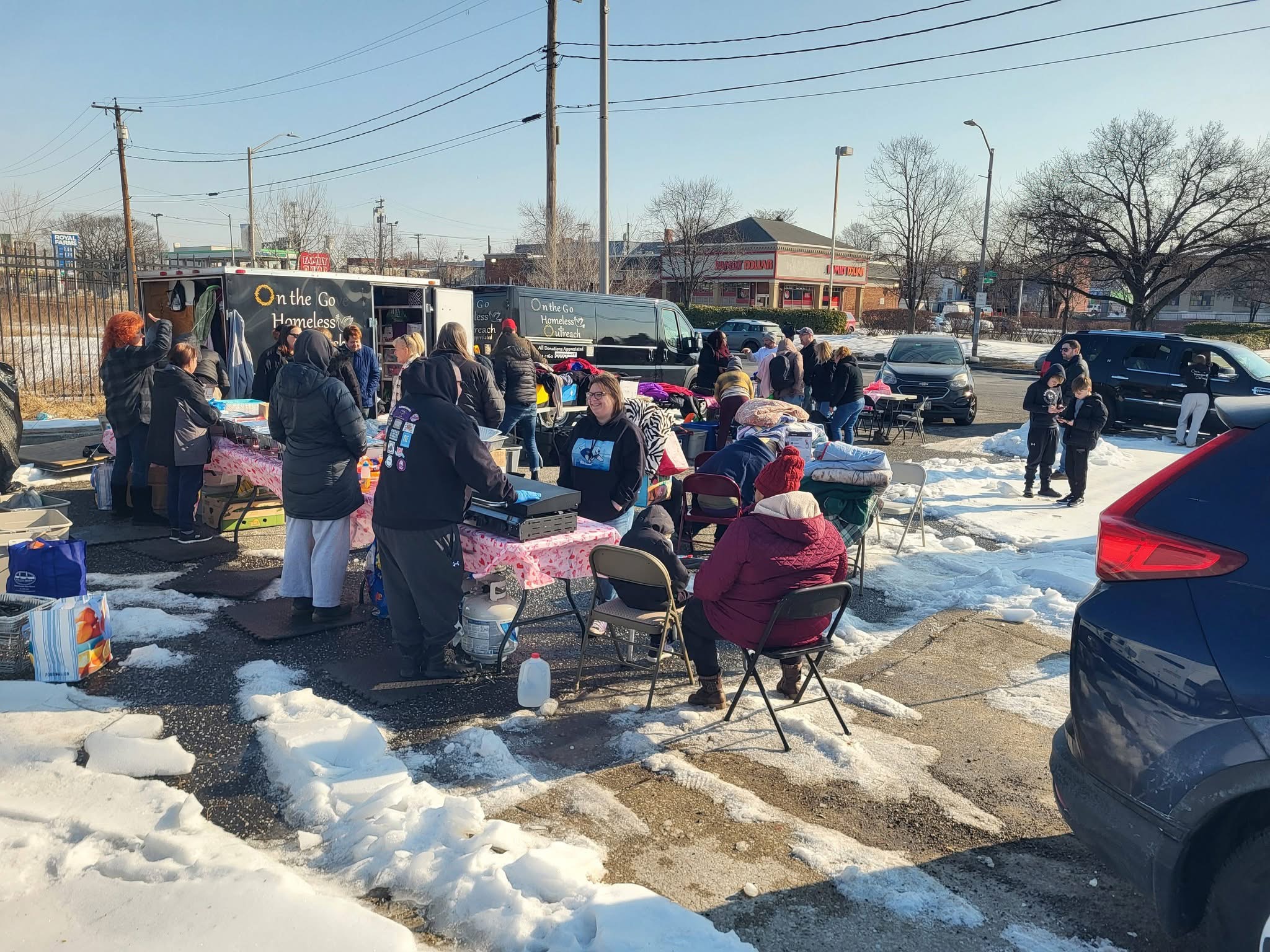 People gather around tables with pink tablecloths outdoors on snow-covered ground, with cars and buses in the background.
