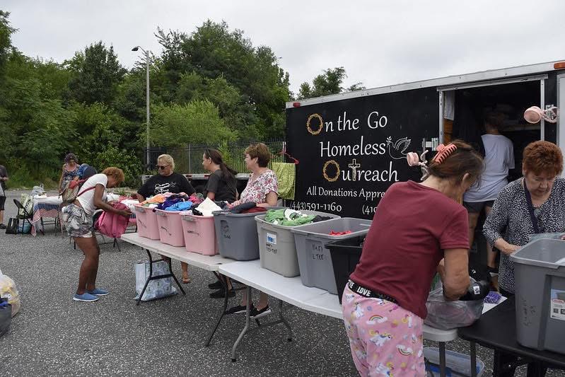 People gather around tables with donation bins outside a mobile outreach vehicle, trees and cloudy sky in background.