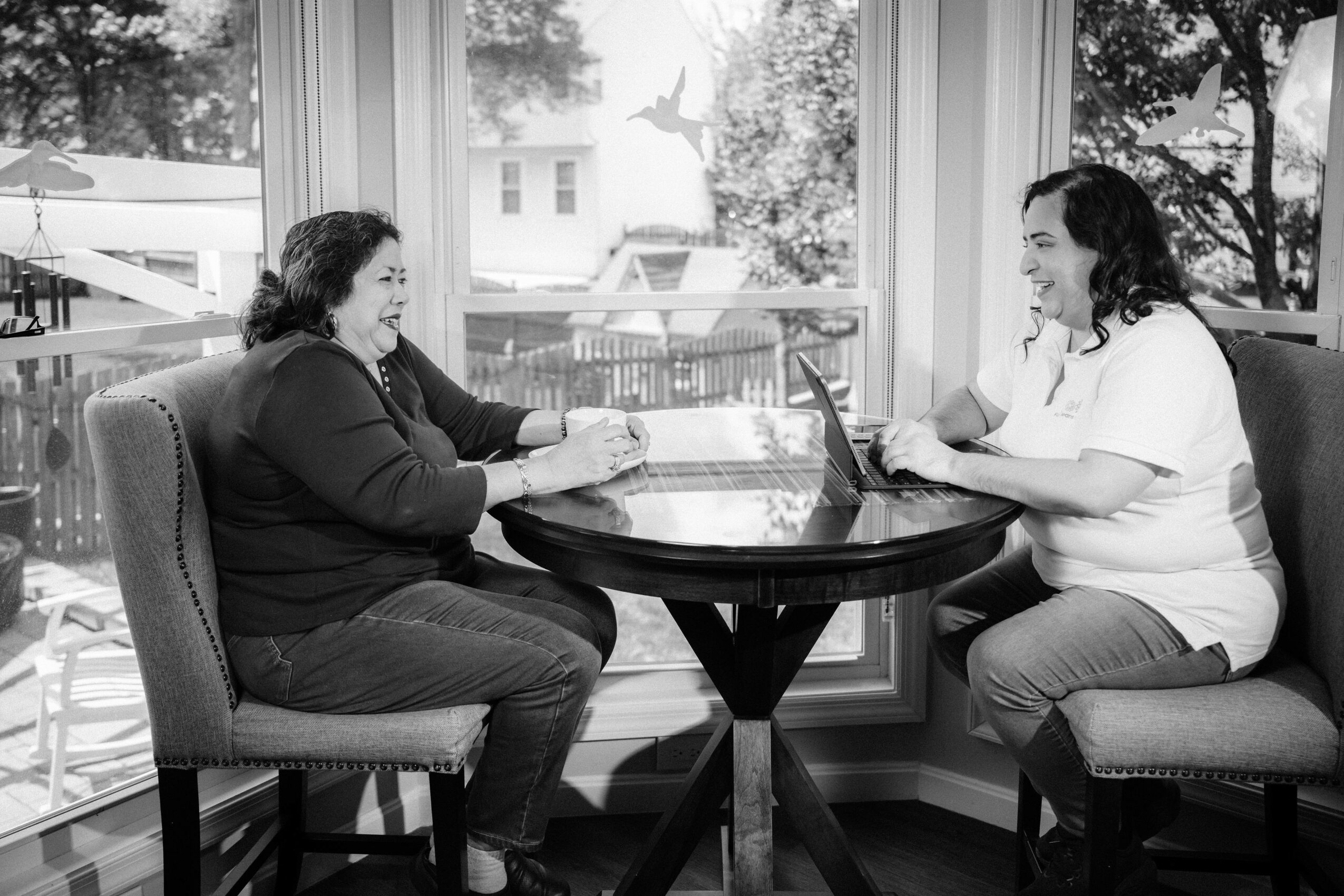 Two women sit at a round table, smiling and talking, with large windows and trees outside in the background.
