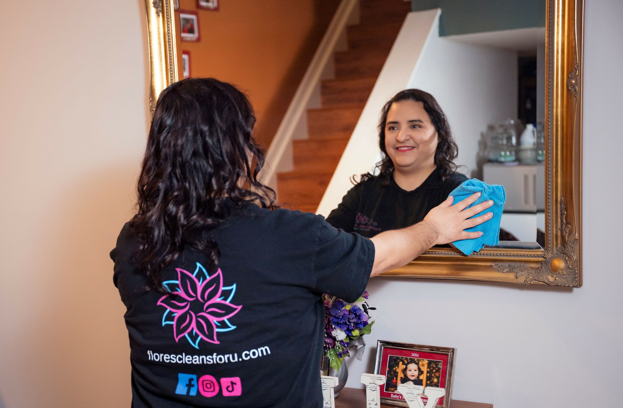 Woman cleaning a mirror with a cloth, smiling at her reflection, in a home interior.